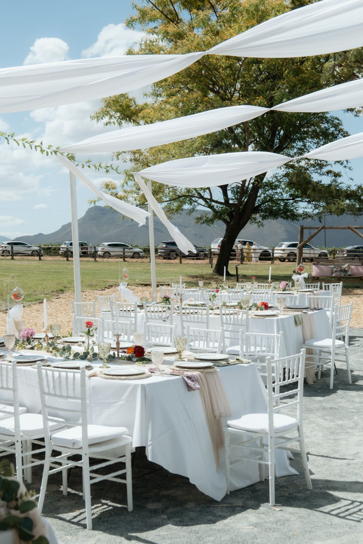 Outdoor wedding reception setup with white draped canopy, round tables, and white chiavari chairs.