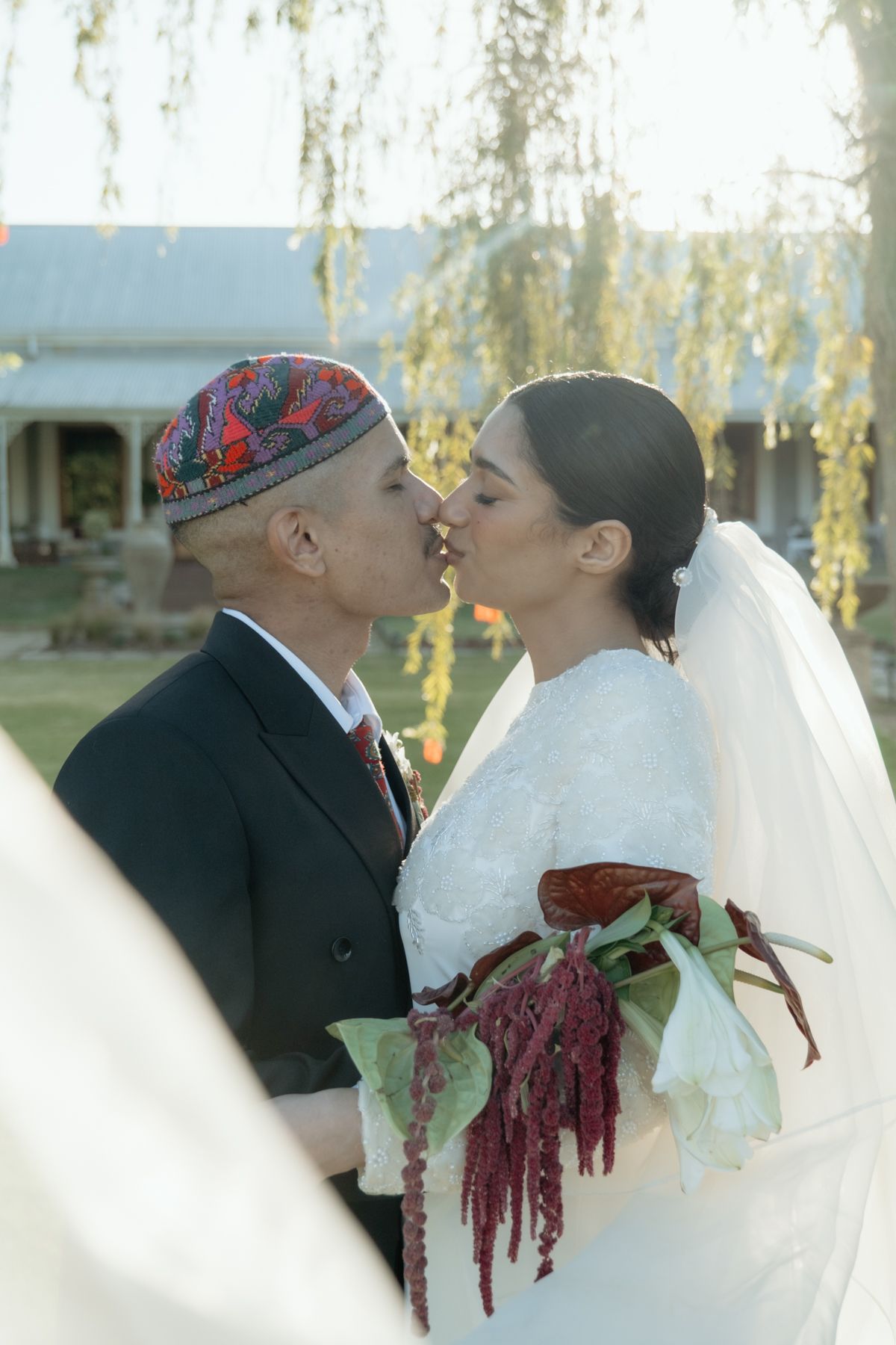 Bride and groom sharing a romantic kiss outdoors, bride holding a lush red and green bouquet.