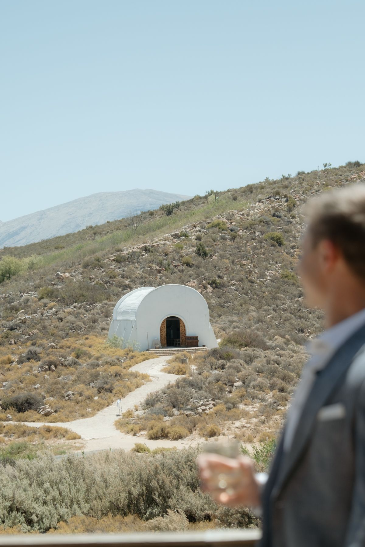 Groom in grey suit holds champagne glass overlooking white dome chapel on arid hillside wedding venue.