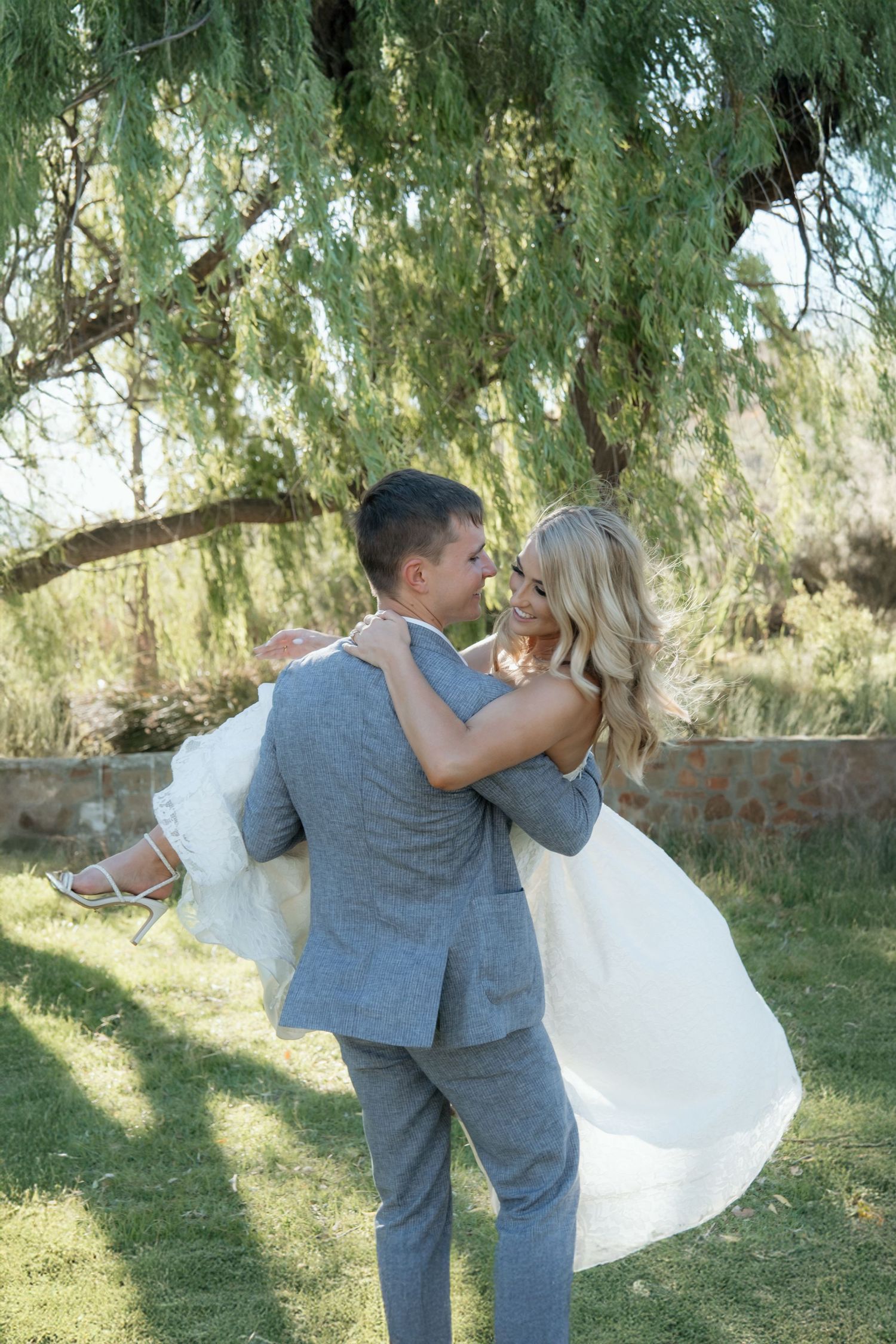 Groom in gray suit lifts bride in white gown under weeping willow tree in sunny outdoor setting.