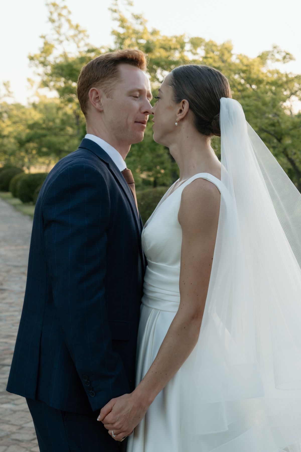 Bride in white gown with veil and groom in navy suit lean in for a kiss while holding hands outdoors.