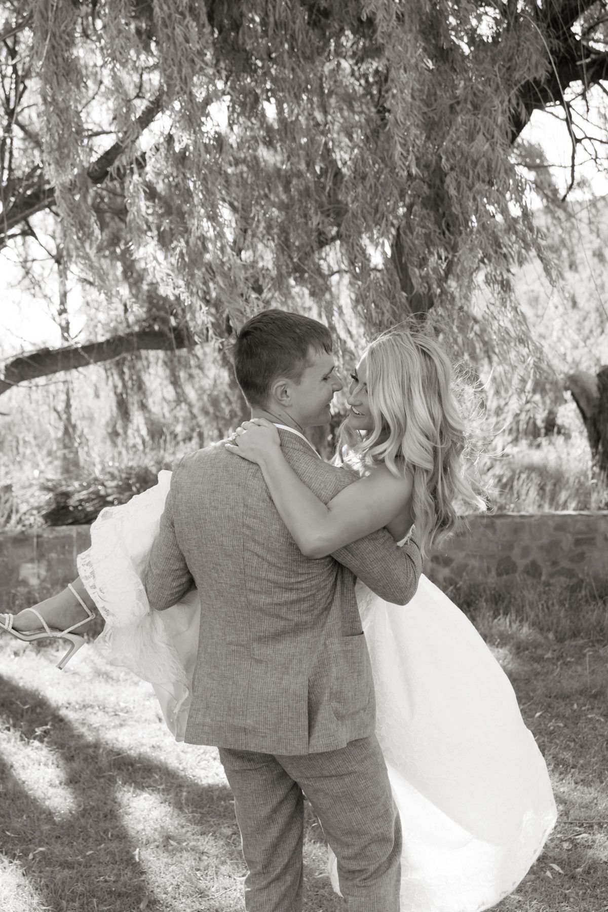 Black and white photo of groom lifting bride in a romantic embrace under a weeping willow tree.