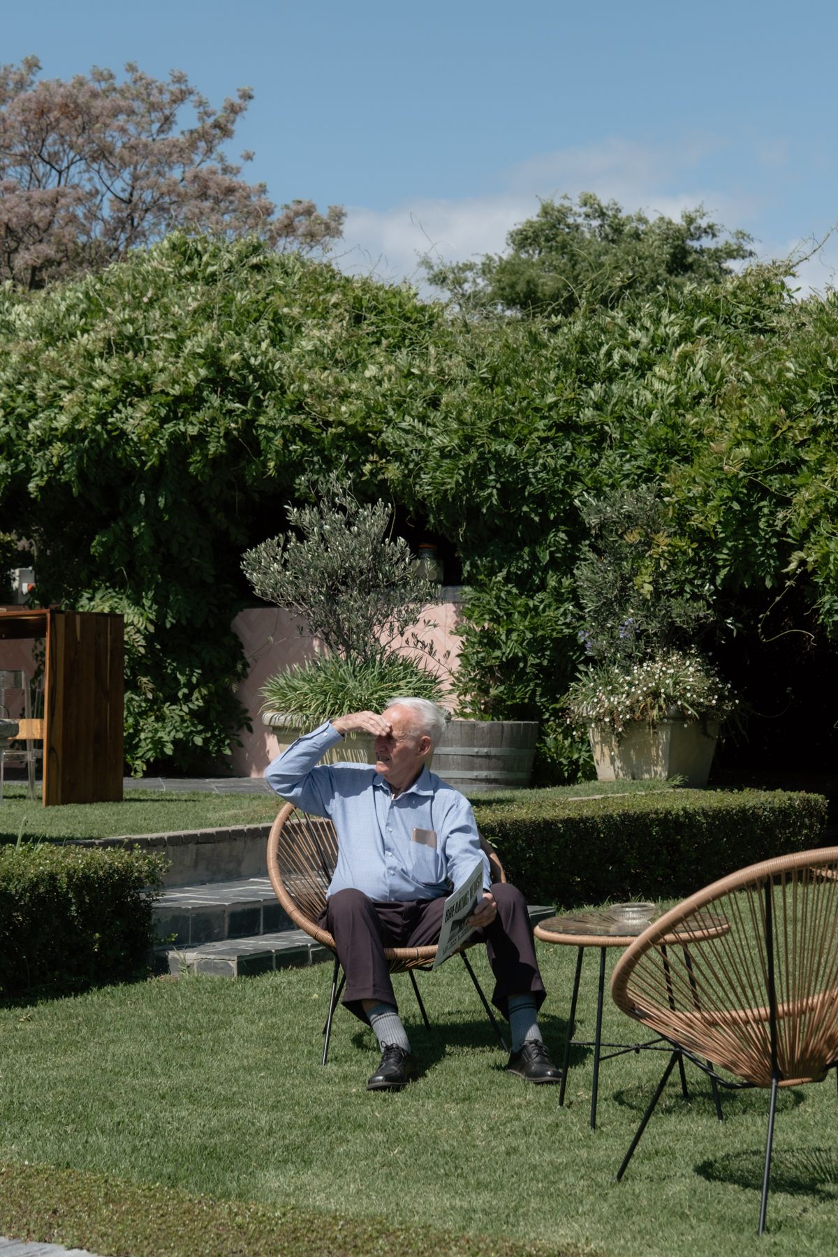 Elderly man relaxes in wicker chair on lush green garden lawn at outdoor wedding.