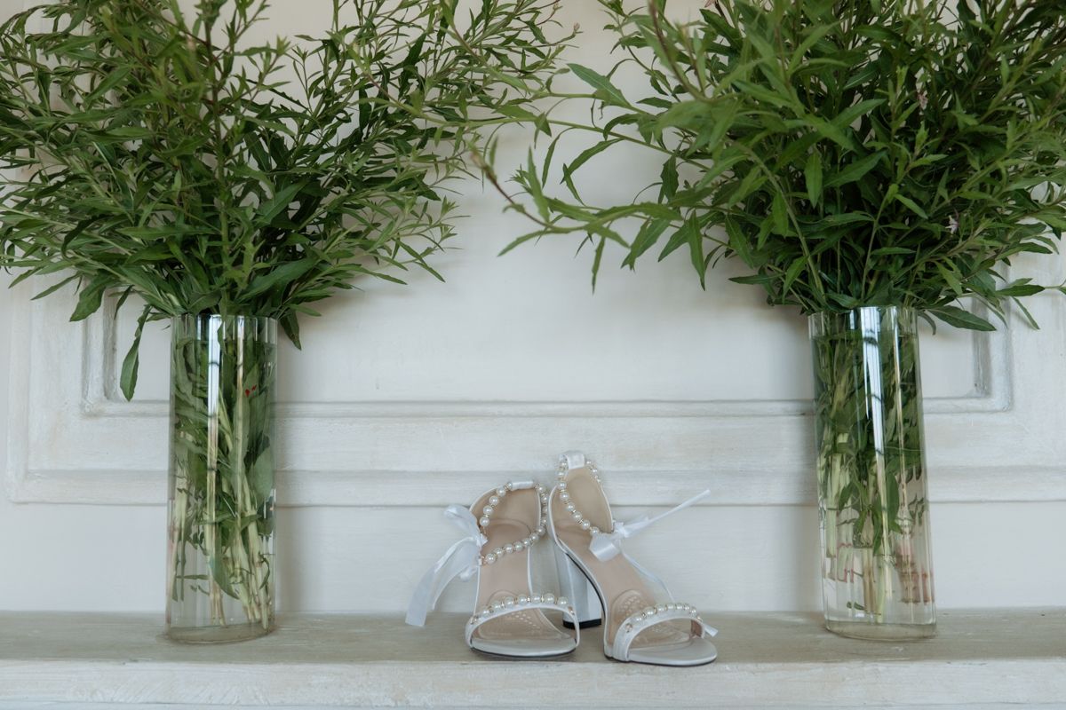 Silver bridal heels displayed between two glass vases with fresh eucalyptus greenery on a white shelf.