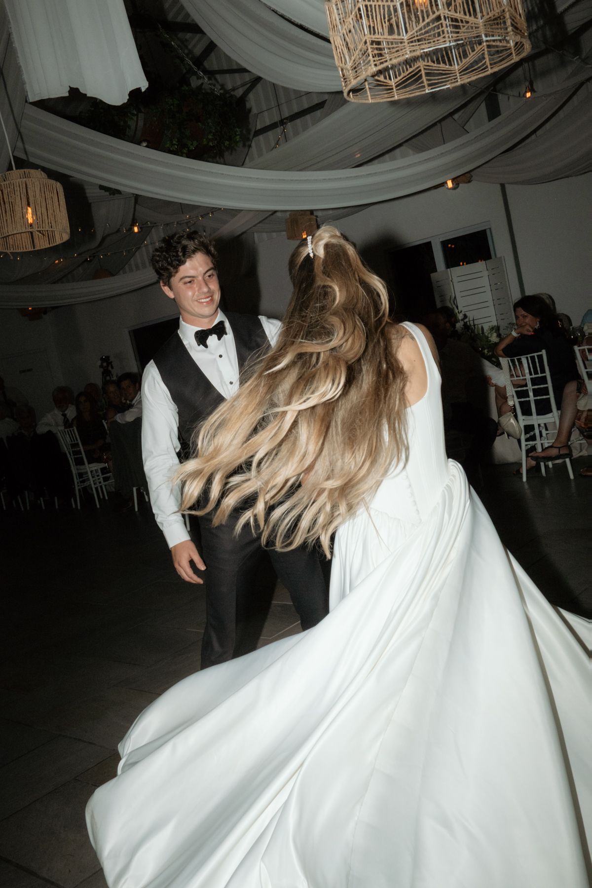 Groom smiling as bride twirls in white wedding gown on dance floor under decorative rattan chandeliers.