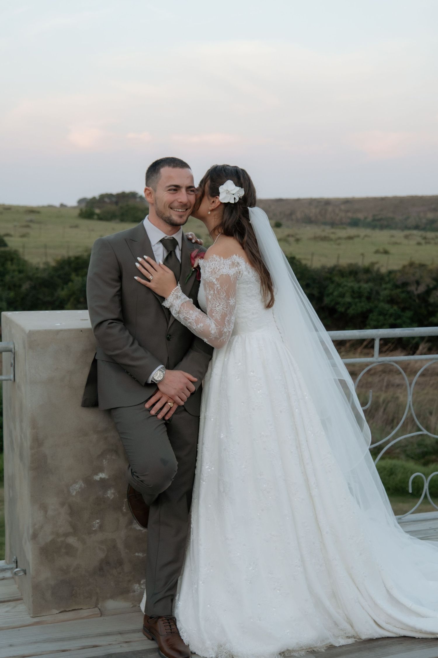 Bride in lace gown kisses groom in olive suit on balcony with scenic green landscape at sunset.