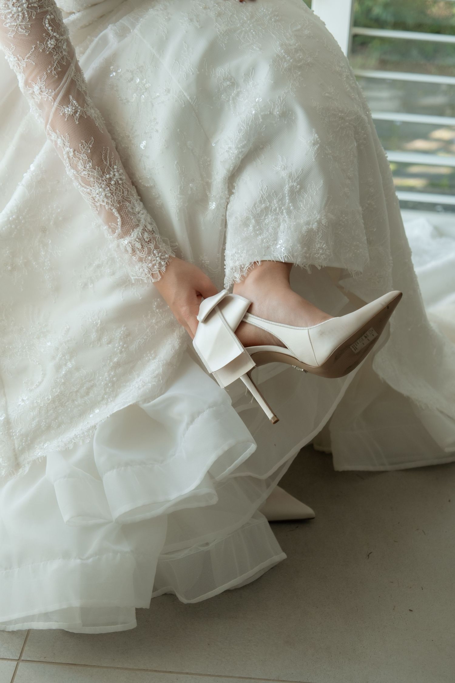 Bride in lace wedding dress putting on white satin heels with bow detail before her wedding ceremony.