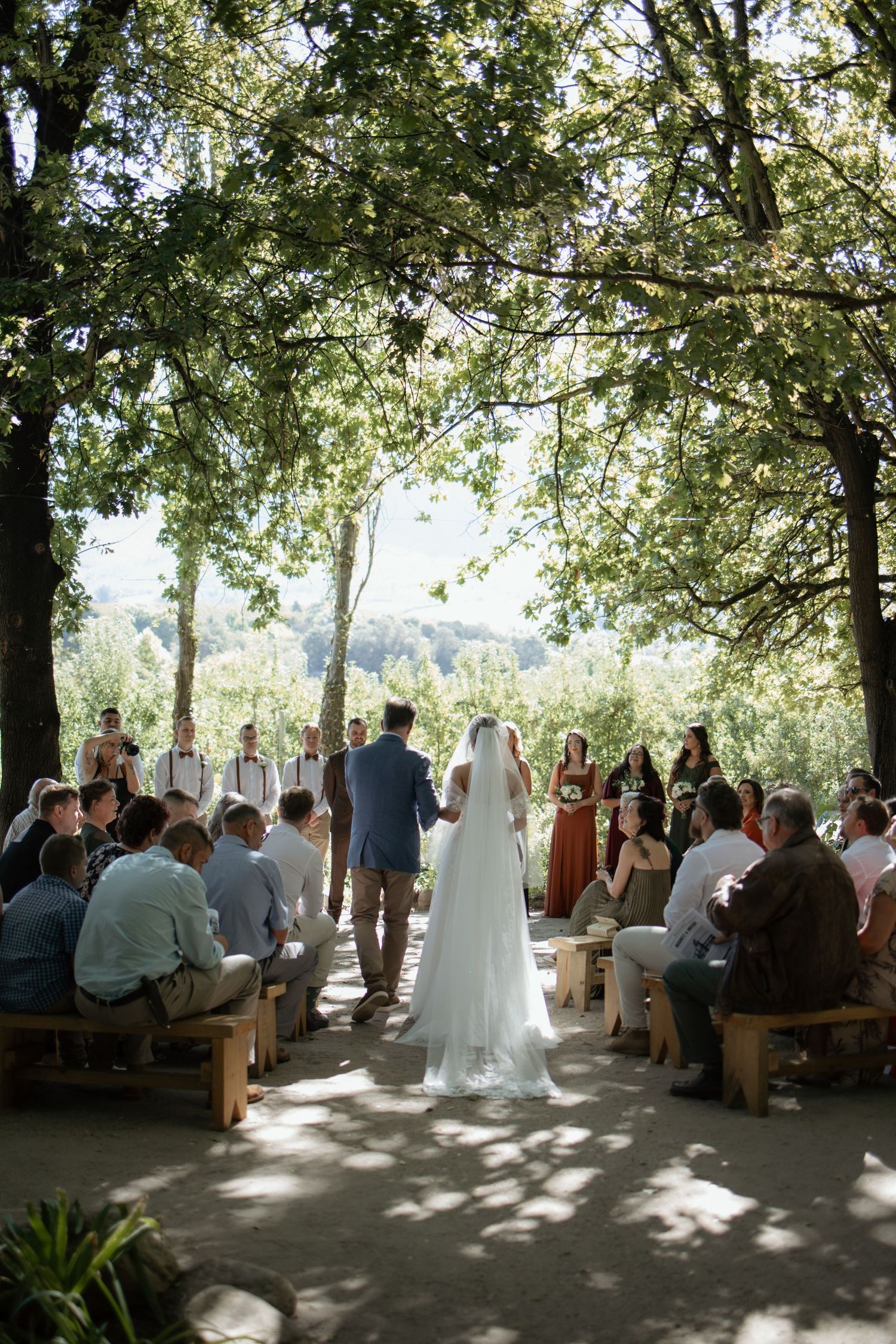 Bride and groom at outdoor wedding ceremony under lush green trees with guests seated on wooden benches.