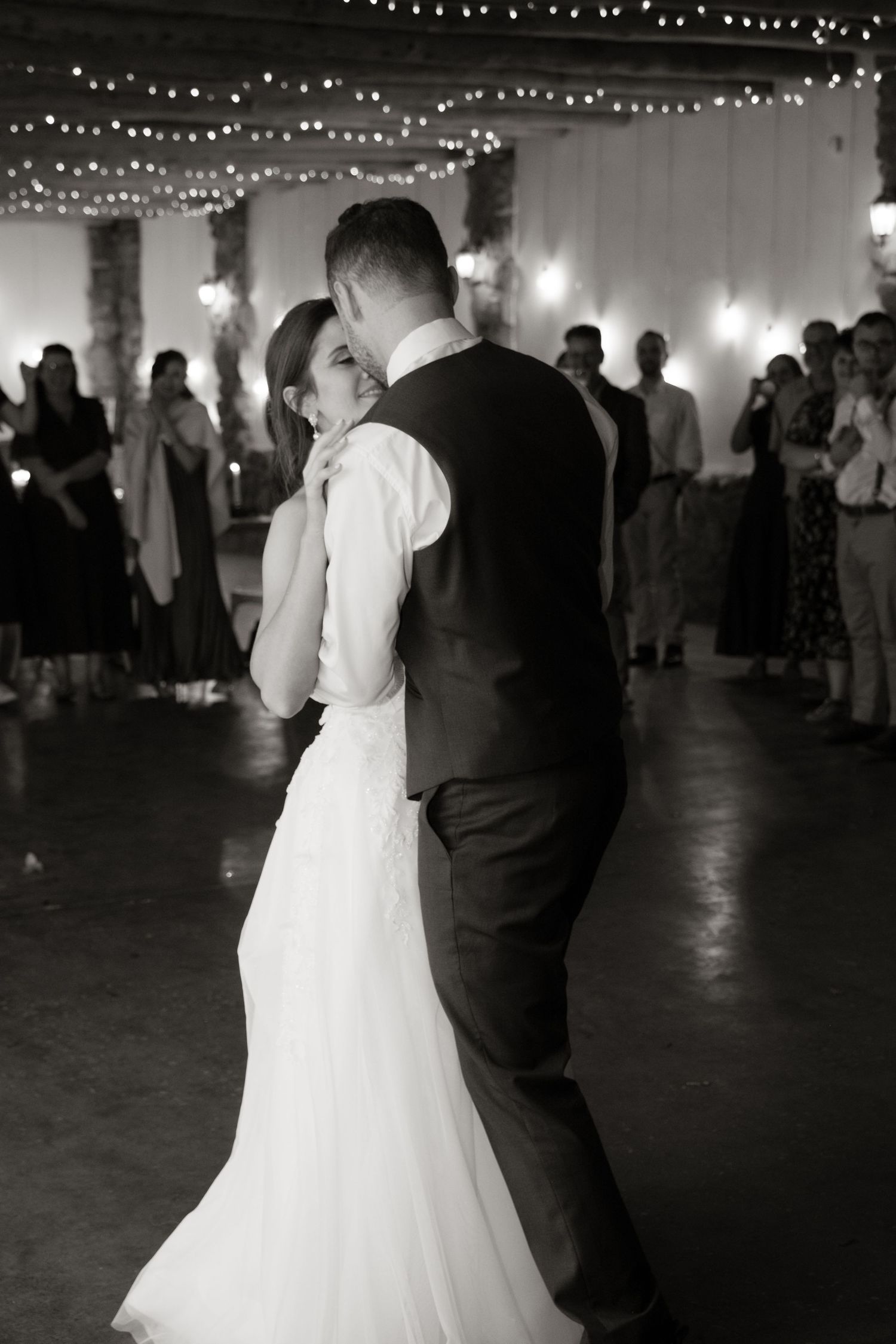 Black and white photo of bride and groom sharing first dance under string lights at wedding reception.