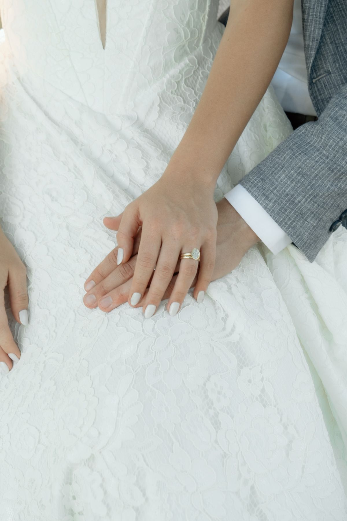 Close-up of bride and groom holding hands showing wedding rings on white lace wedding dress.