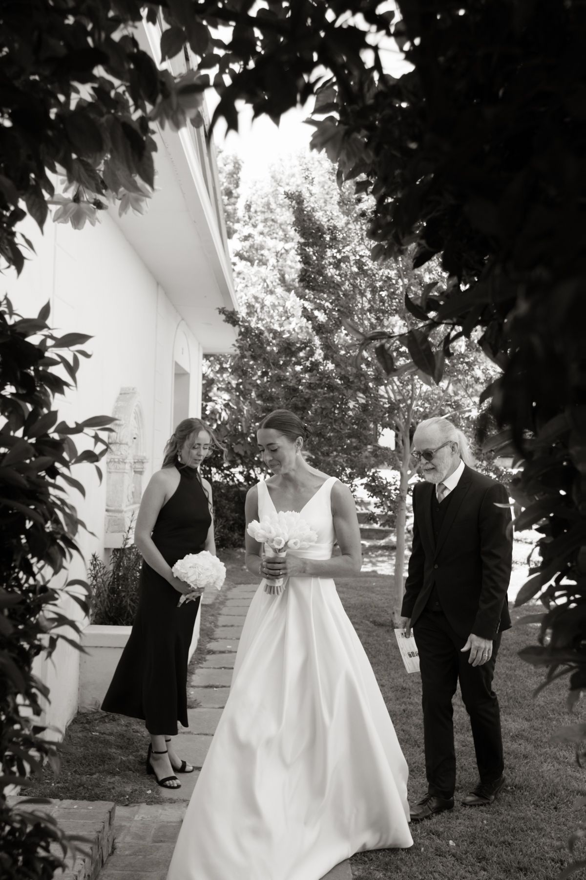 Black and white photo of a bride in a white gown walking with two guests through a garden path.
