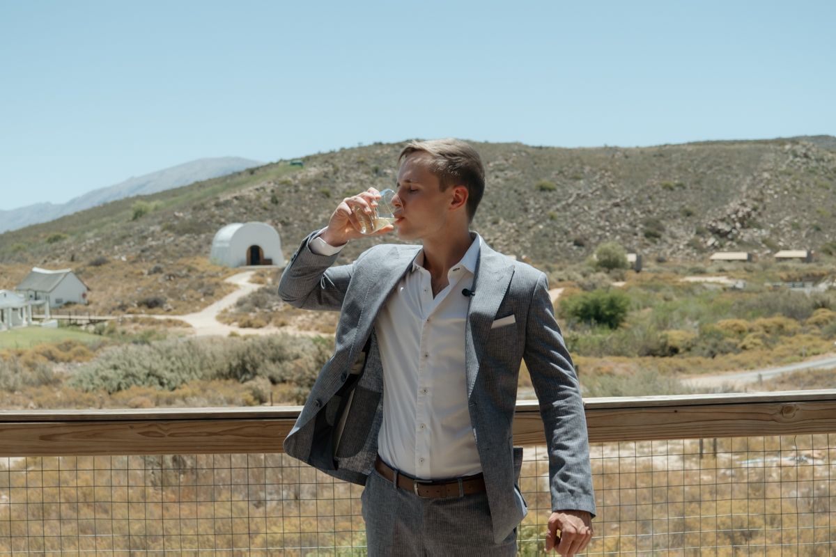 A man in a grey suit drinks wine on a balcony with arid hills in the background.