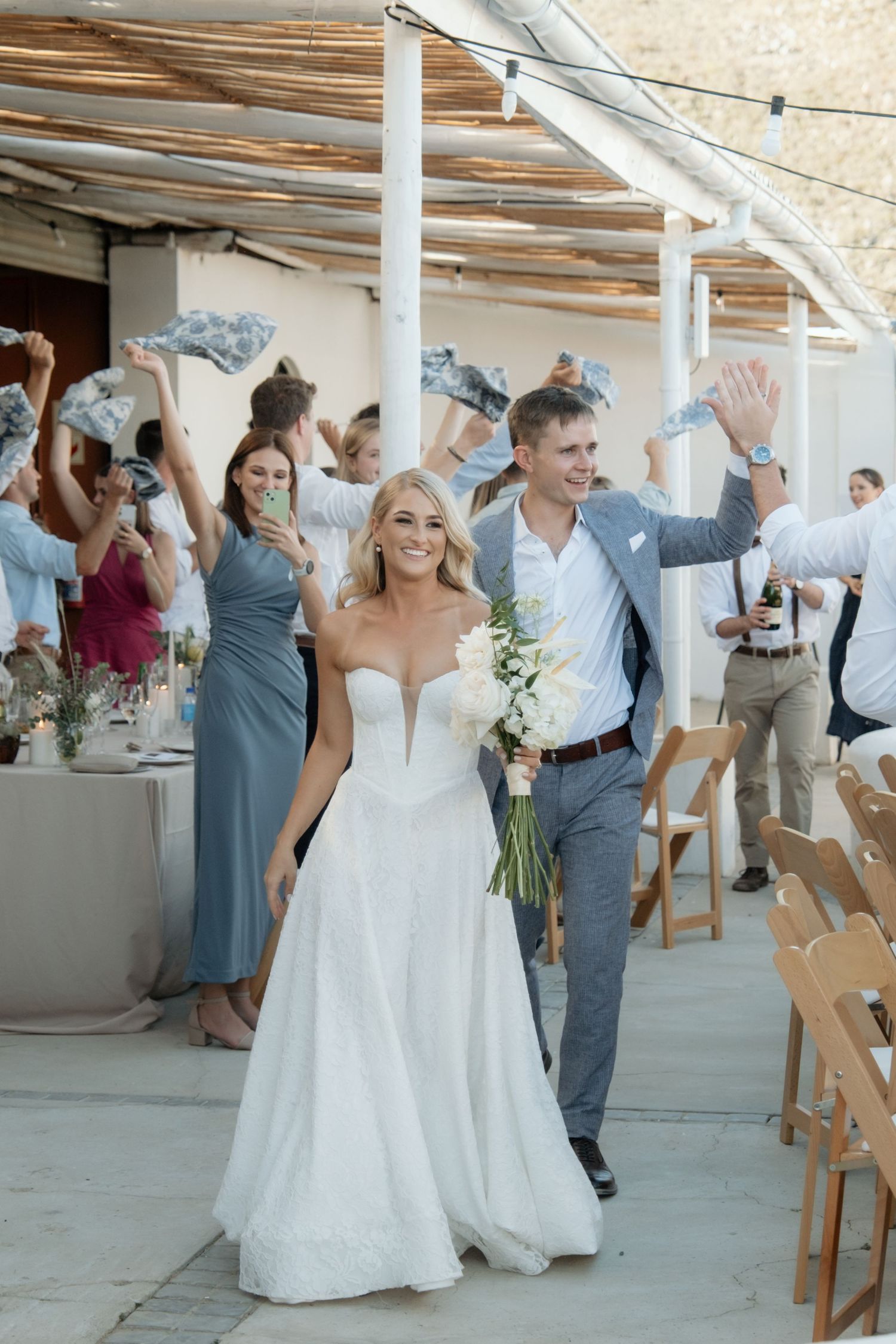 Joyful bride and groom walk through celebrating guests waving at their outdoor wedding reception ceremony exit.