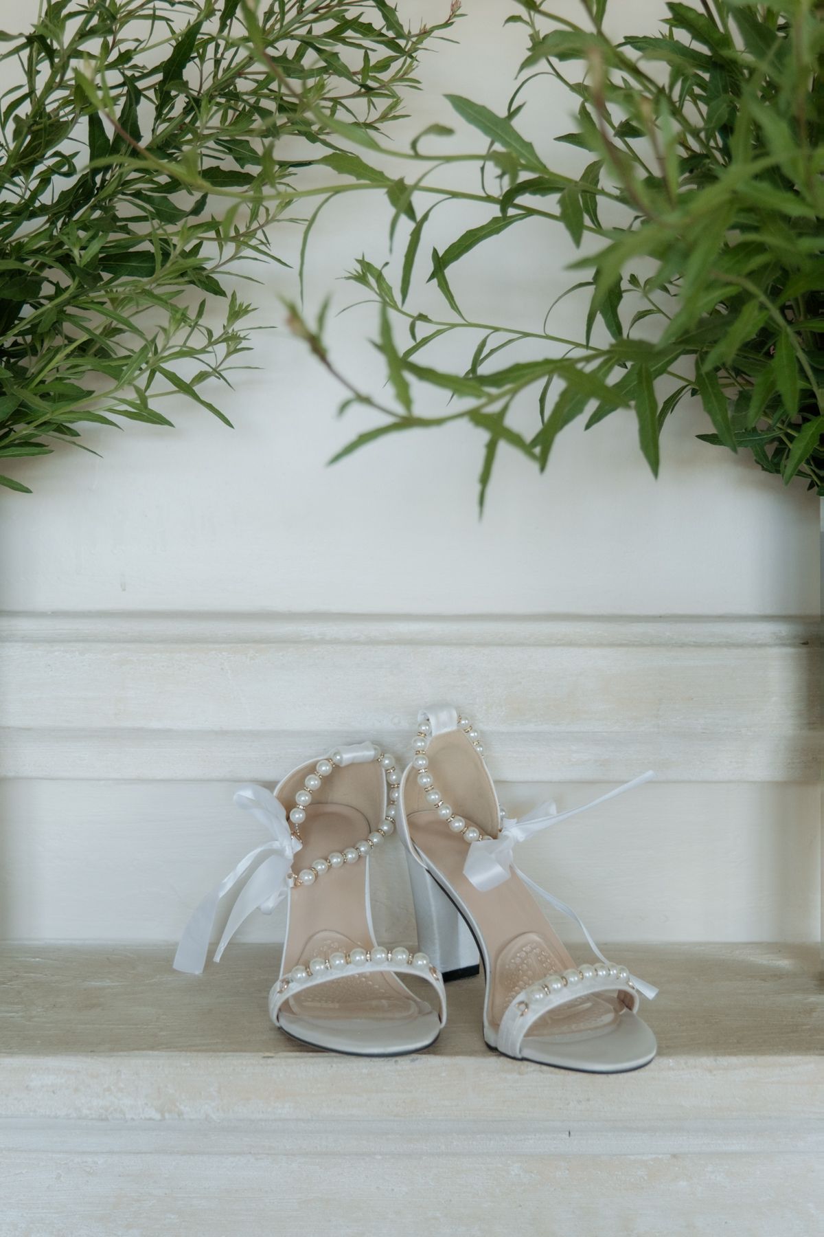 Elegant white bridal heels adorned with pearl details and ribbon ties displayed on a marble surface with greenery.