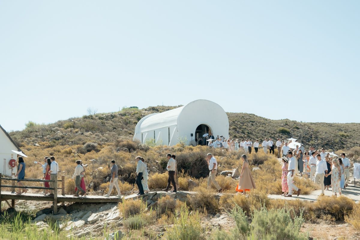 Wedding guests gather outside a white arched venue on a sunny hillside landscape.