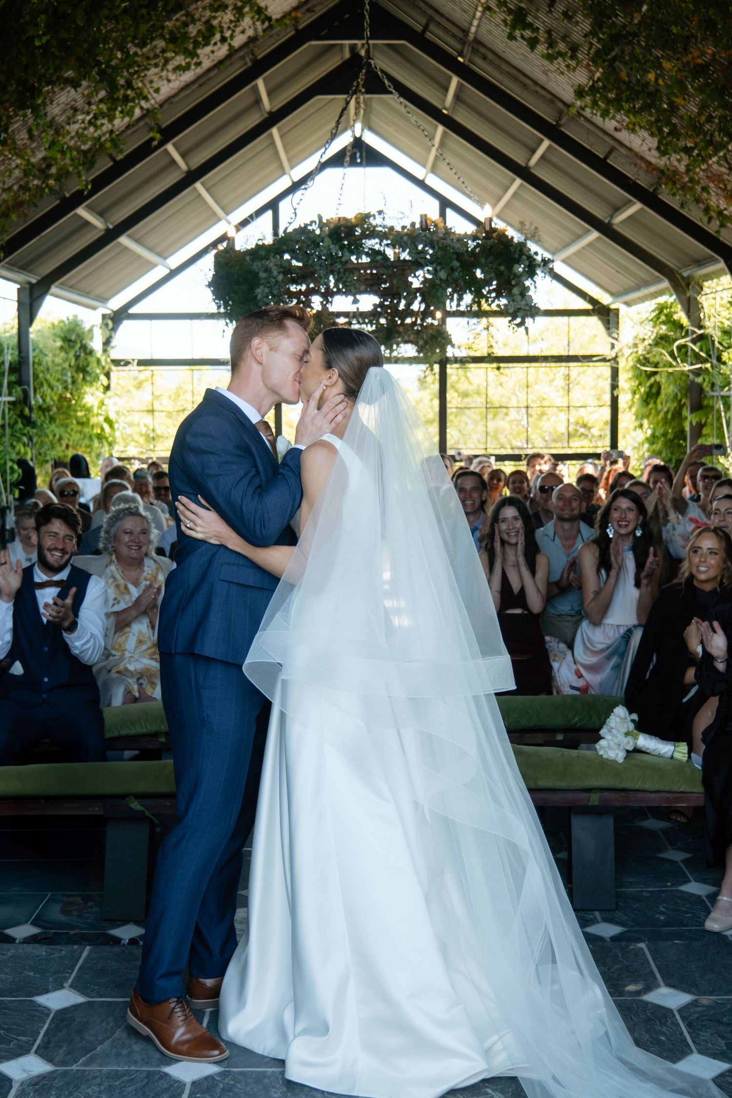 Bride and groom sharing first kiss at altar in glass chapel surrounded by cheering wedding guests.