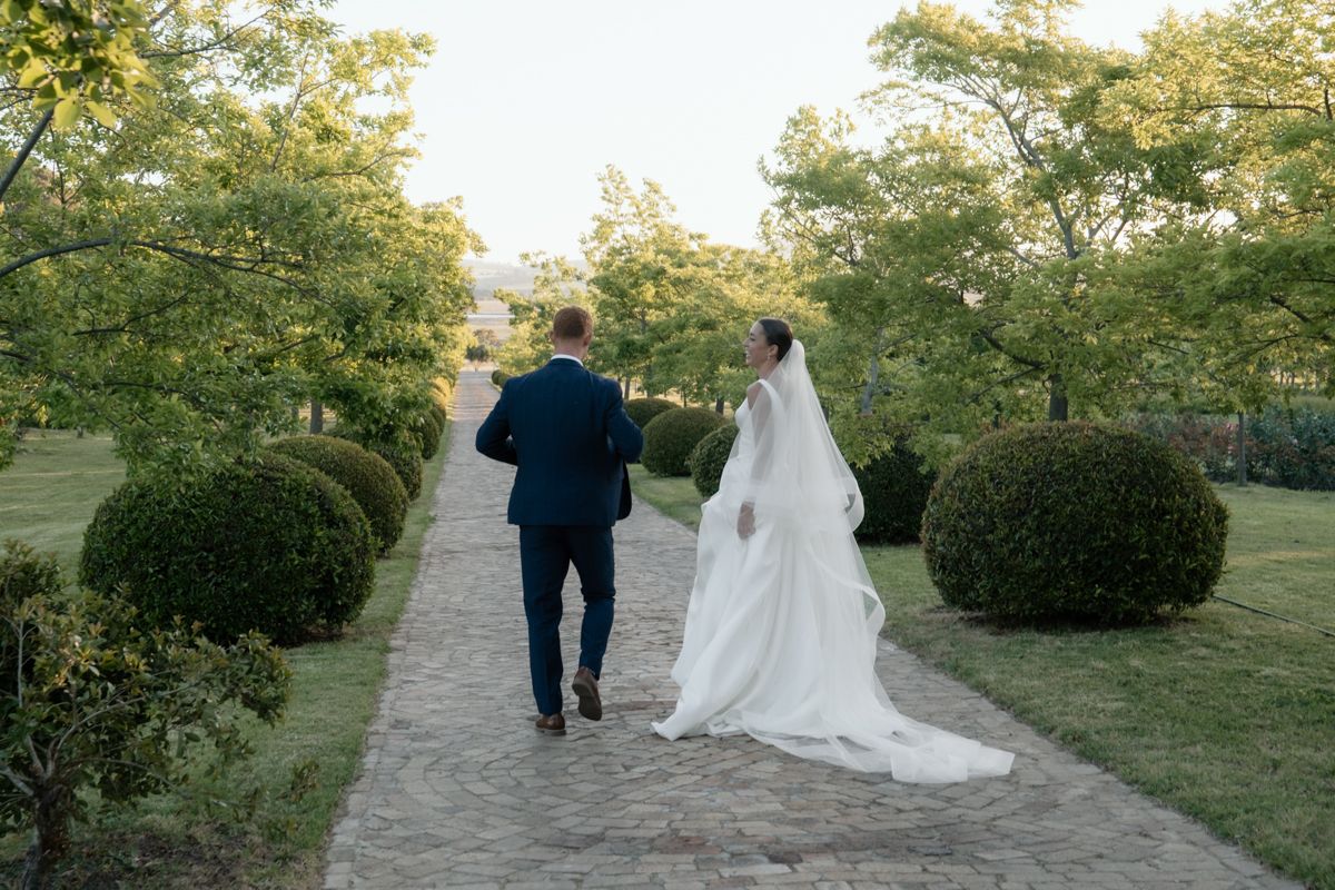 Bride and groom walk hand in hand along a garden path surrounded by lush greenery on their wedding day.