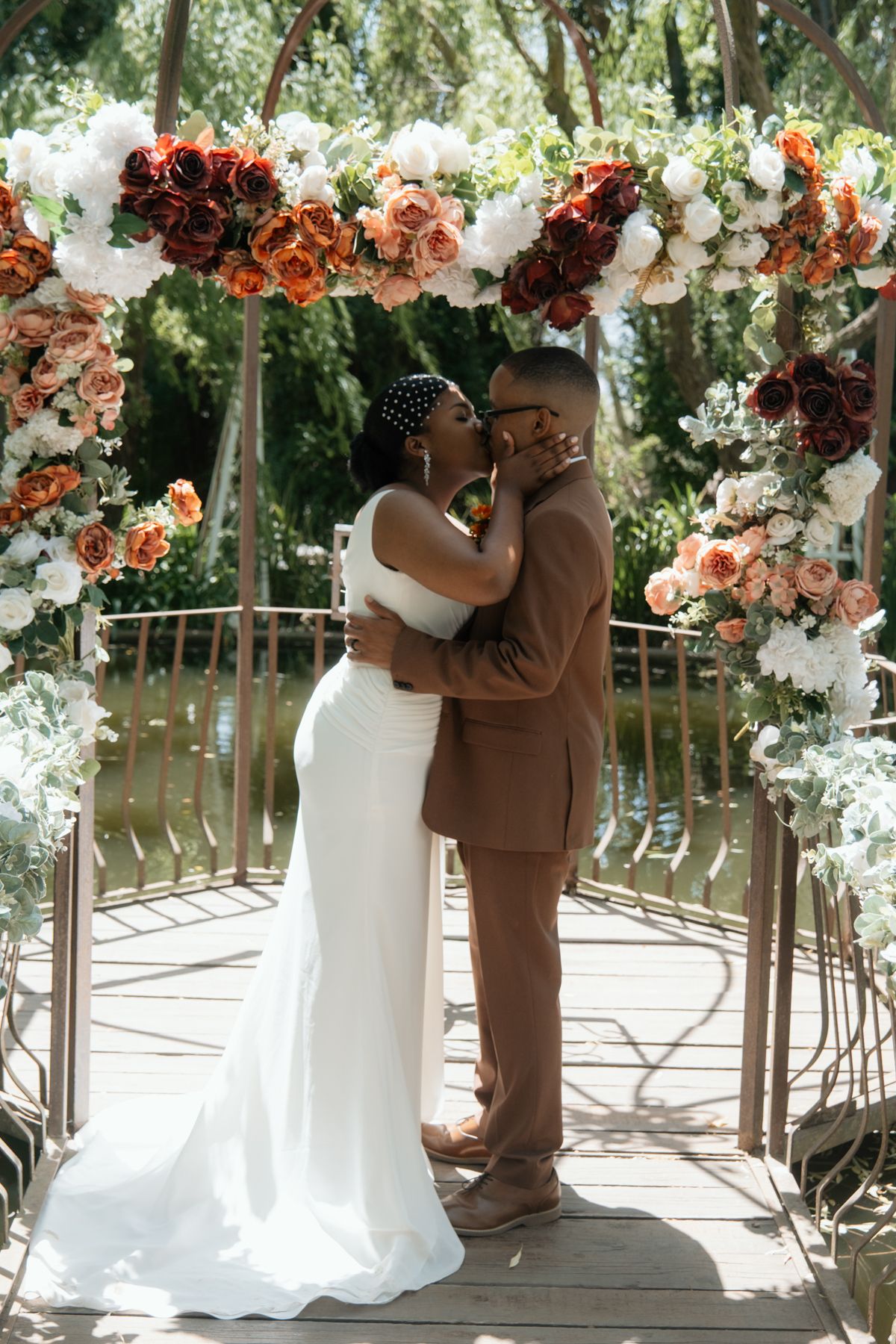 A Black bride and groom share a kiss under a floral arch adorned with red, orange, and white roses at their wedding ceremony.