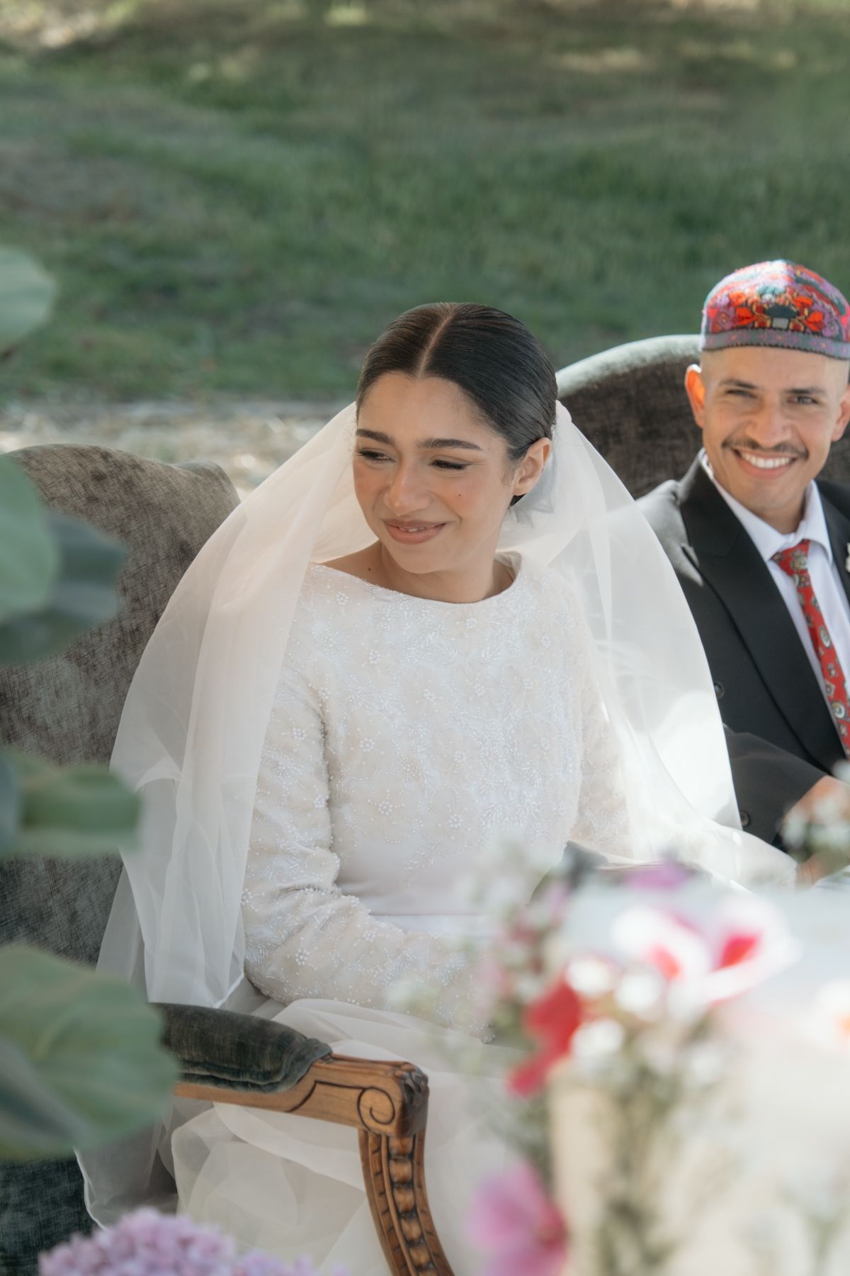Bride in white gown and veil smiling during outdoor wedding ceremony, groom in embroidered cap behind her.