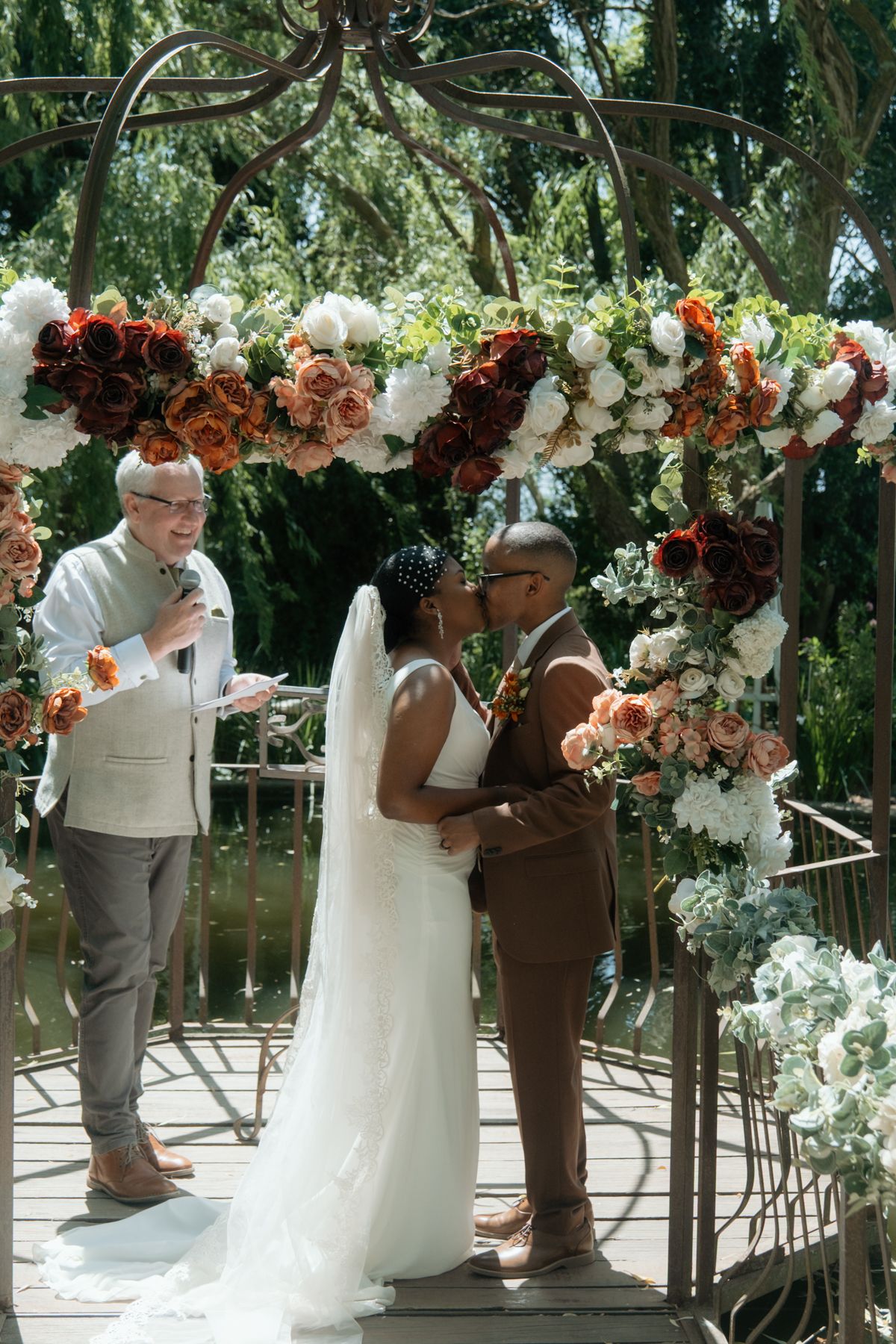 Couple shares first kiss under ornate floral arch with white and orange roses as officiant reads ceremony.