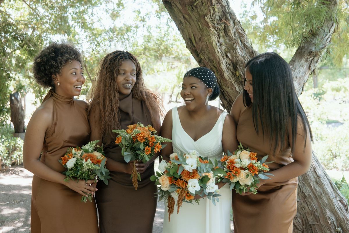 Bride in white dress and bridesmaids in brown gowns share a joyful laugh while holding colorful bouquets outdoors.