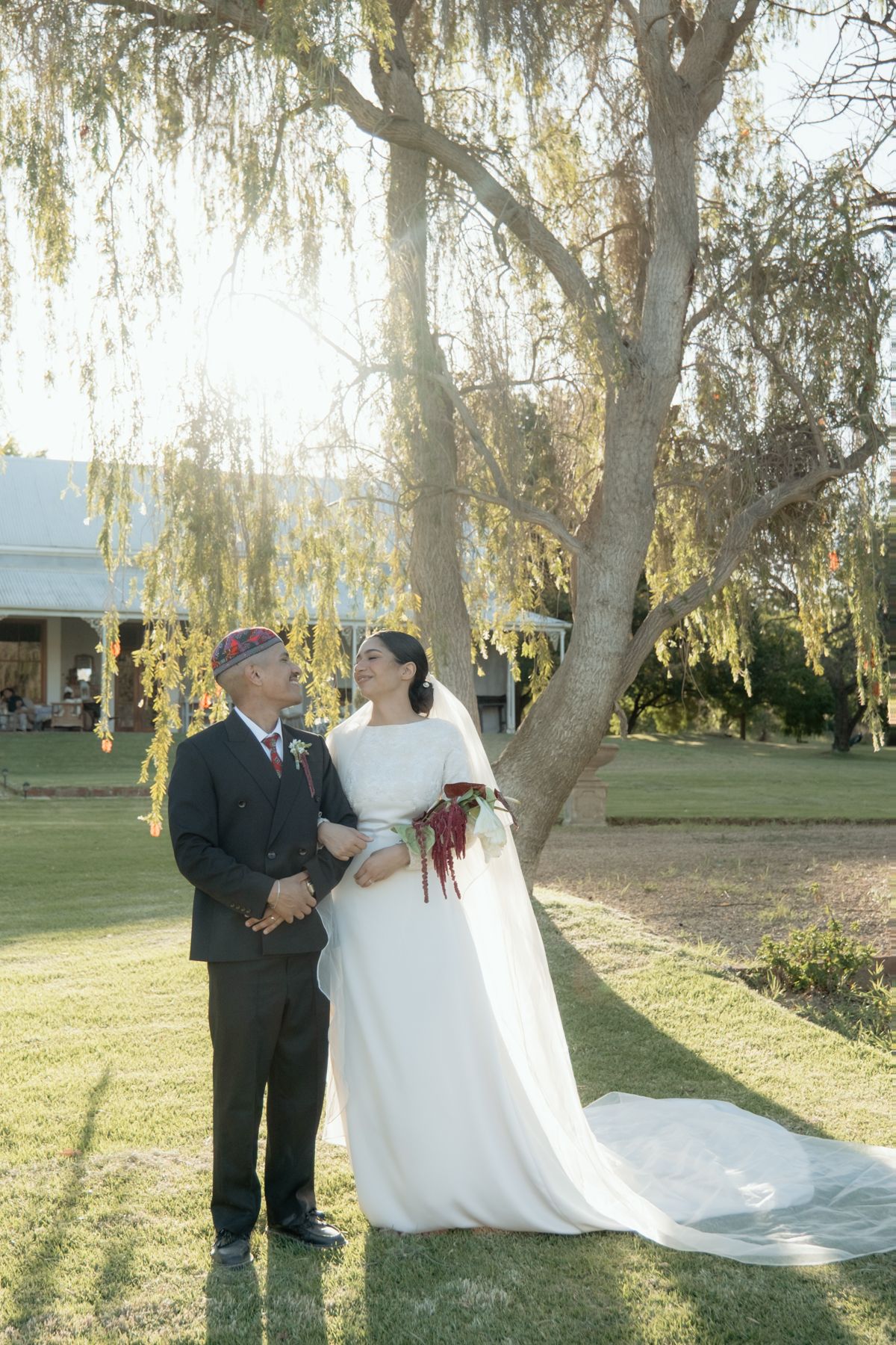 Wedding couple standing under a sunlit willow tree, bride holding a red amaranth bouquet outdoors.