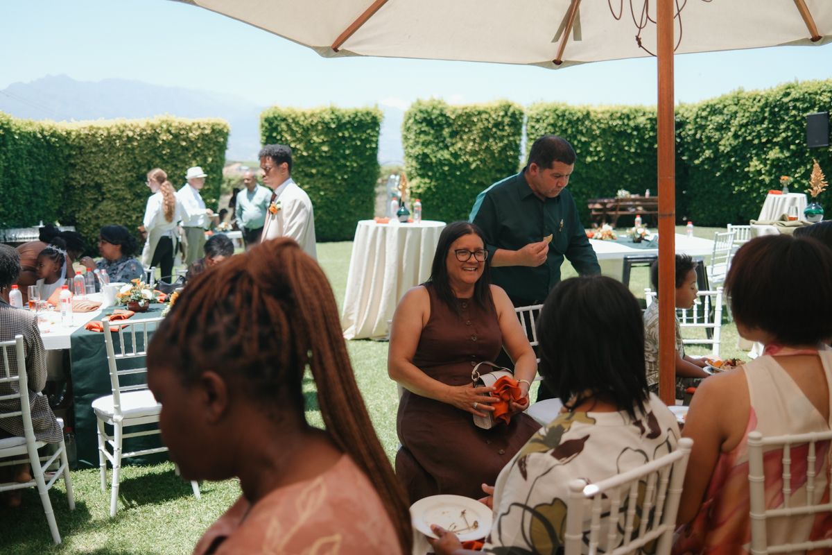 Outdoor wedding reception with guests seated at white chairs under umbrellas on a sunny day.