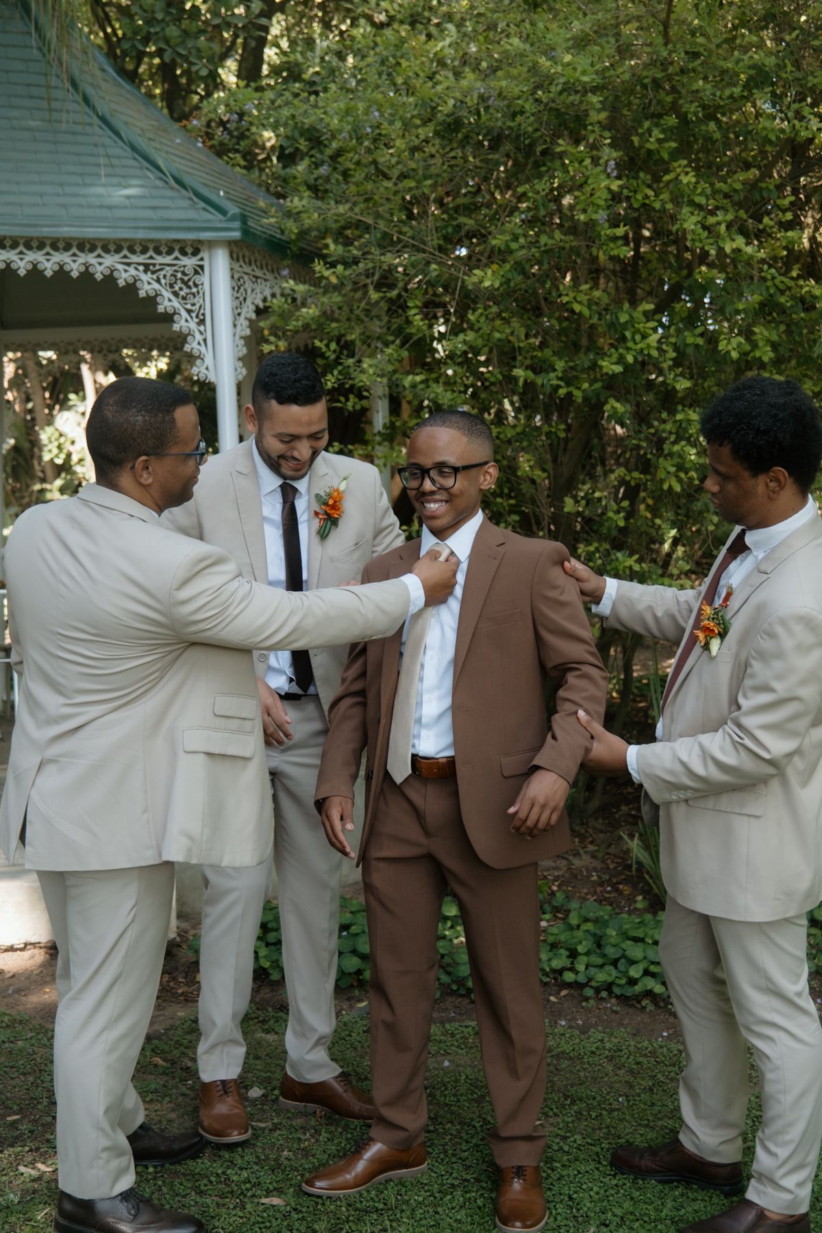 Groomsmen in beige suits help groom in brown suit with tie near elegant garden gazebo.