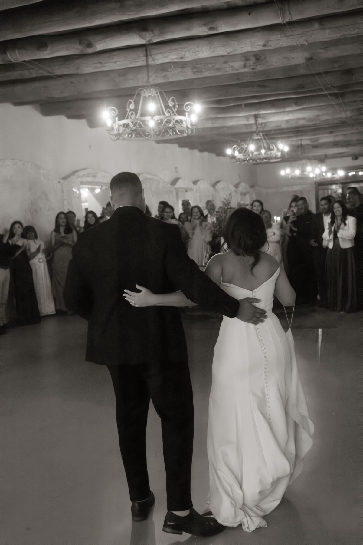 Black and white photo of bride and groom sharing first dance before guests at wedding reception.