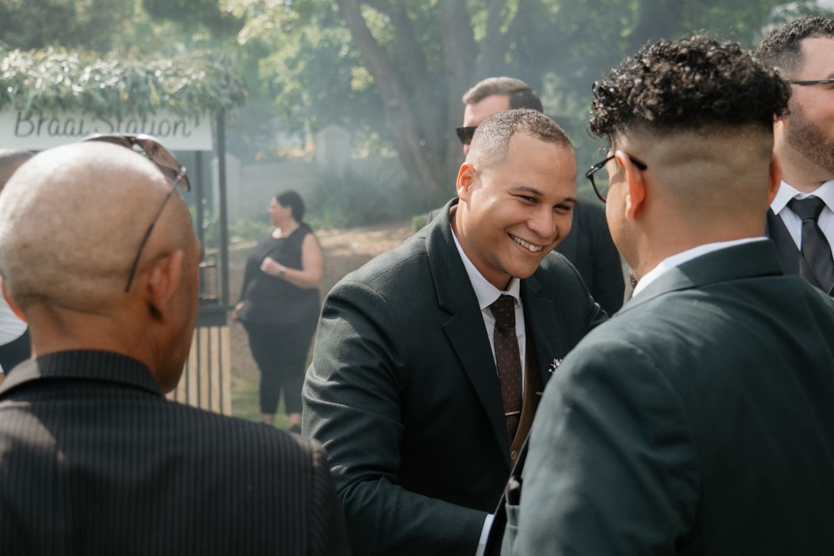 Smiling groom in dark suit greets and shakes hands with wedding guests at outdoor reception.