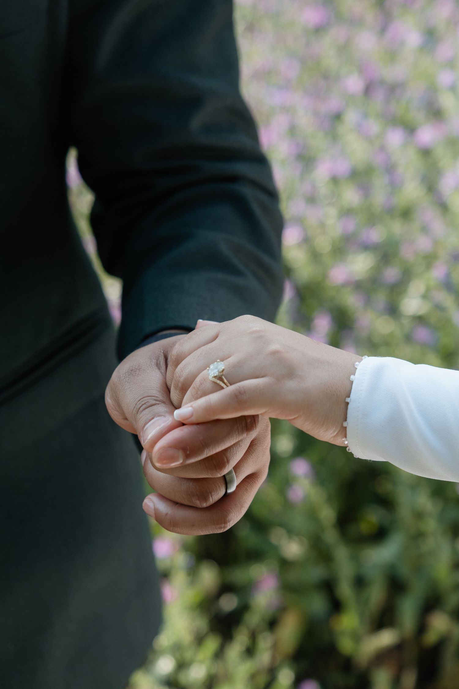 Close-up of bride and groom holding hands, showcasing elegant diamond engagement ring with floral background.