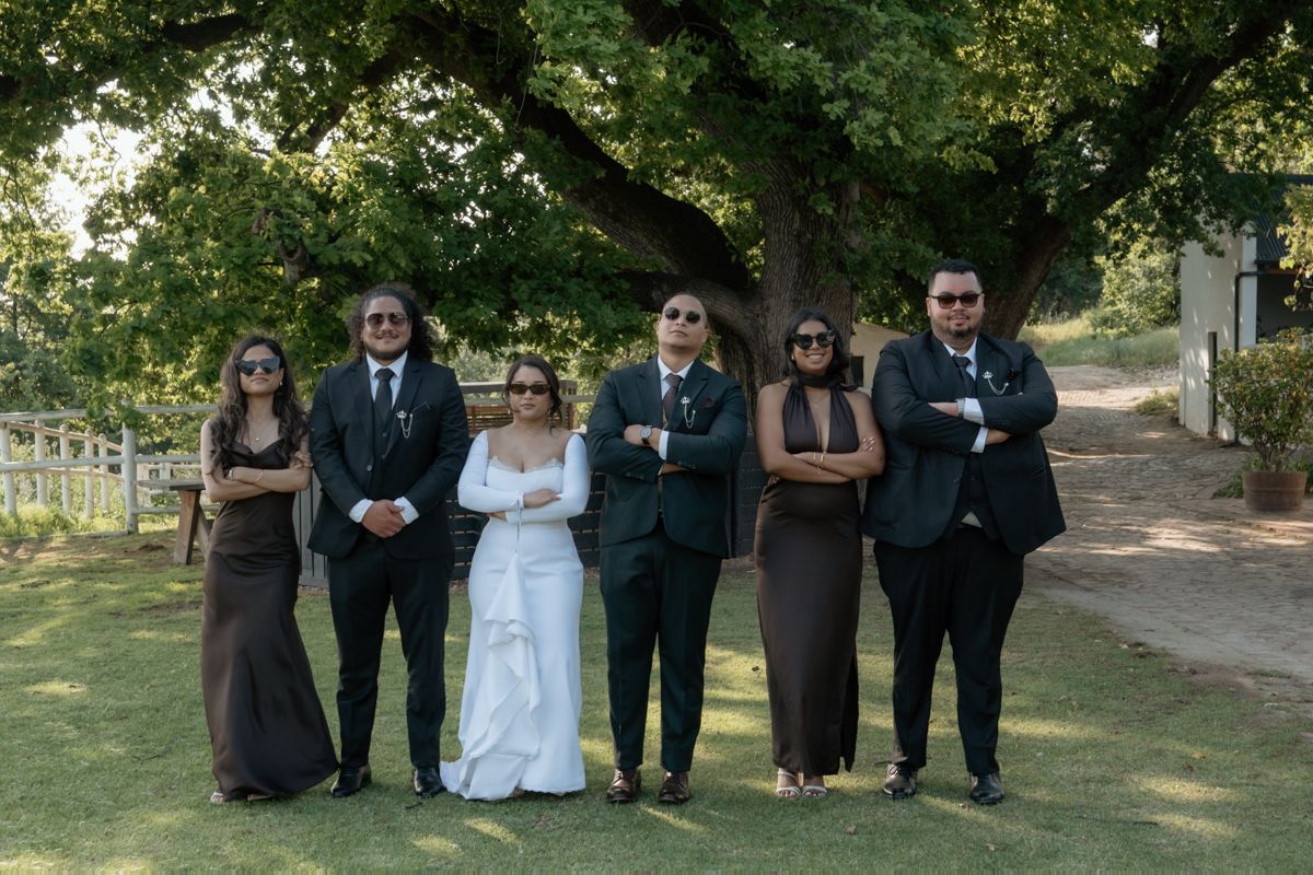 A wedding party of six poses under a large tree, with the bride in white and guests in dark formal attire.