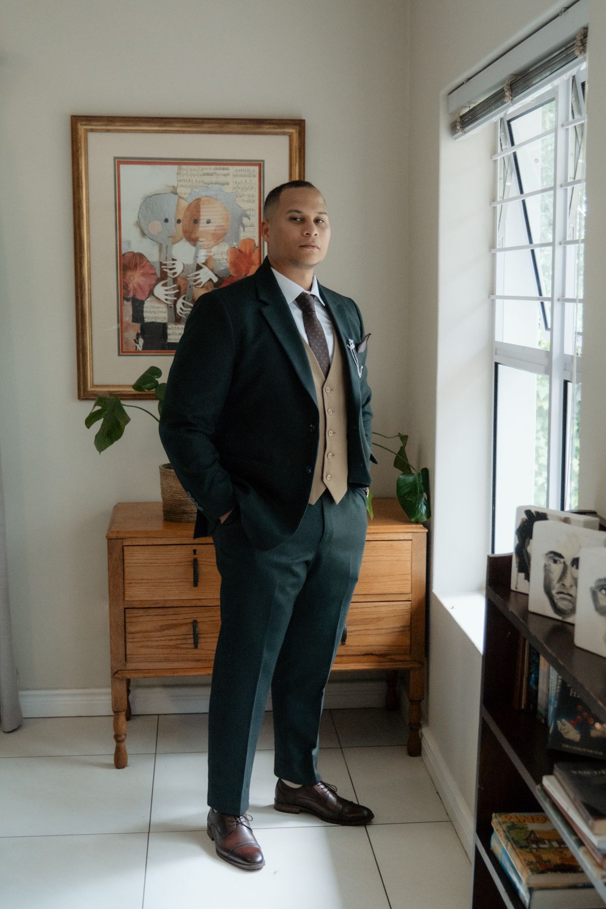 Groom in dark green suit with tan vest poses confidently indoors before wedding ceremony.