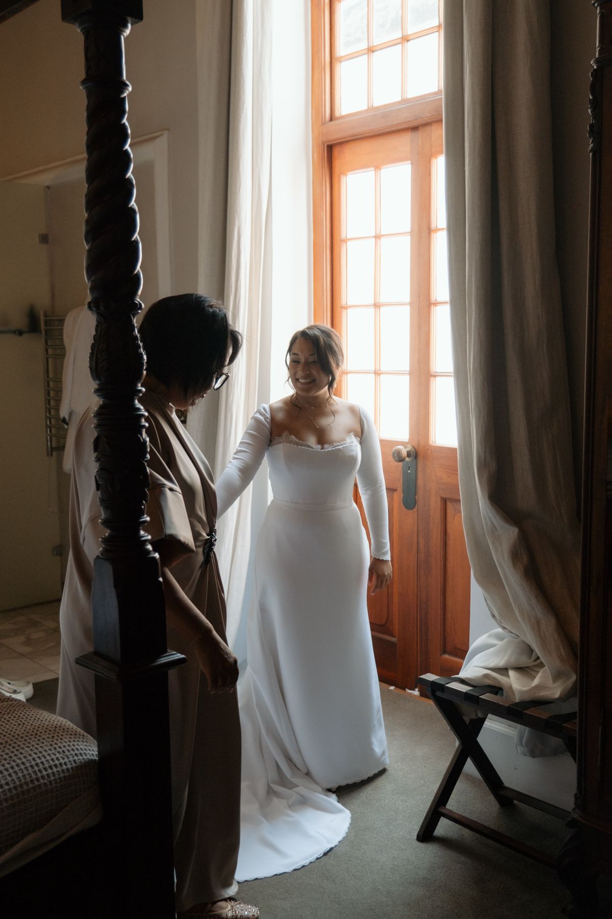 Bride in elegant white gown with gloves stands by sunlit window as bridesmaid helps her prepare.