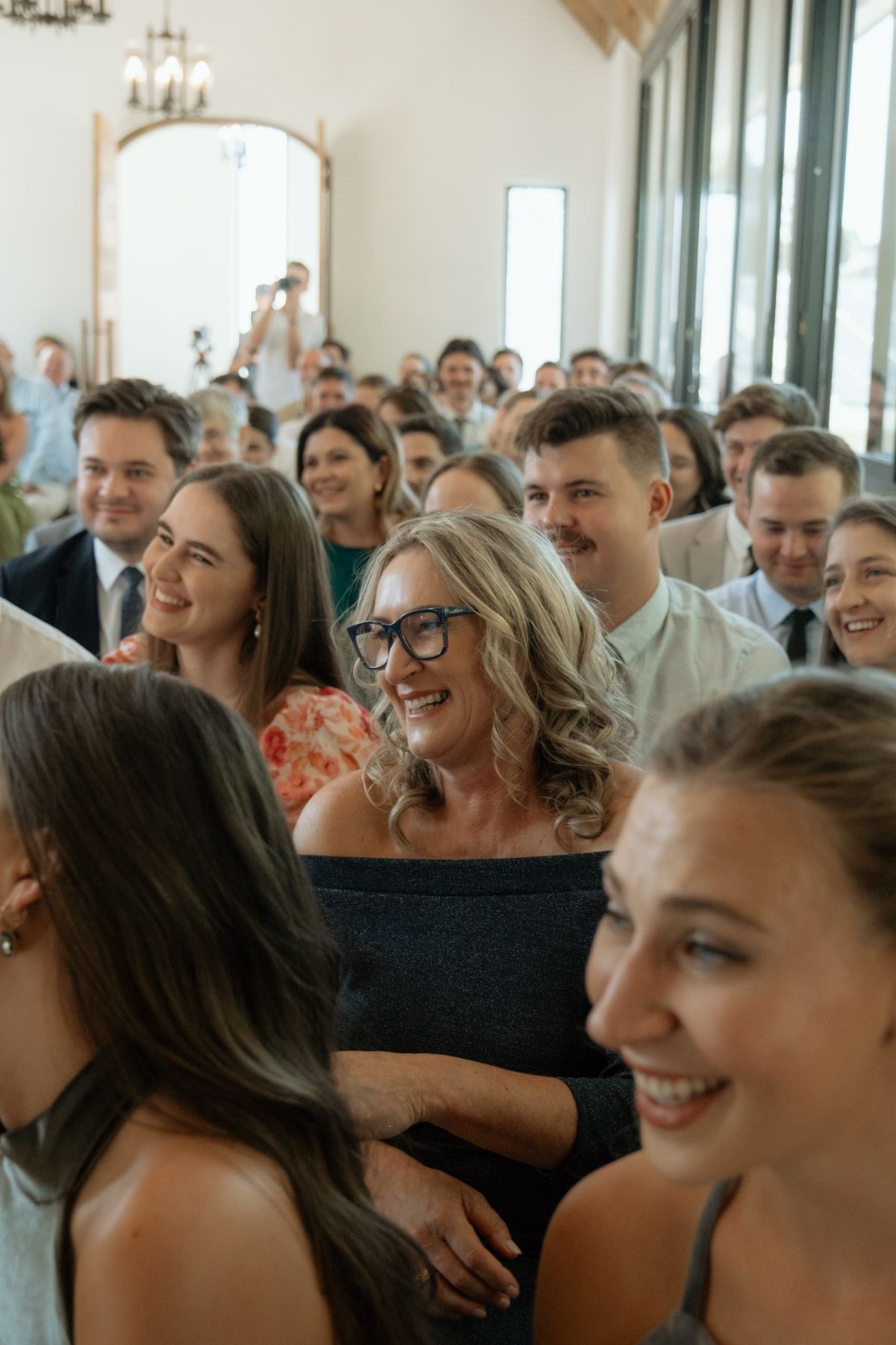 Smiling wedding guests stand and watch the ceremony in a bright white venue.
