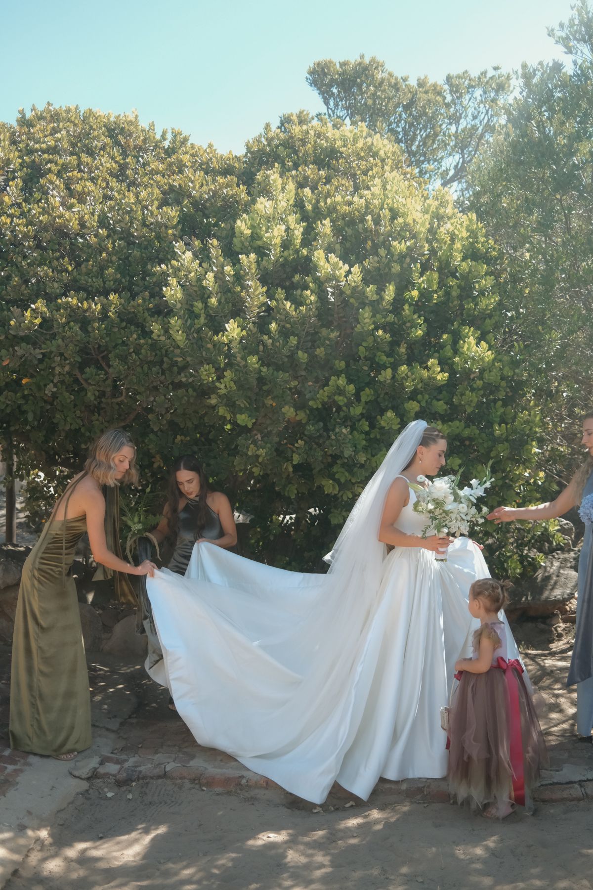 Bridesmaids help a bride with her white gown and veil outdoors, as a flower girl watches nearby.