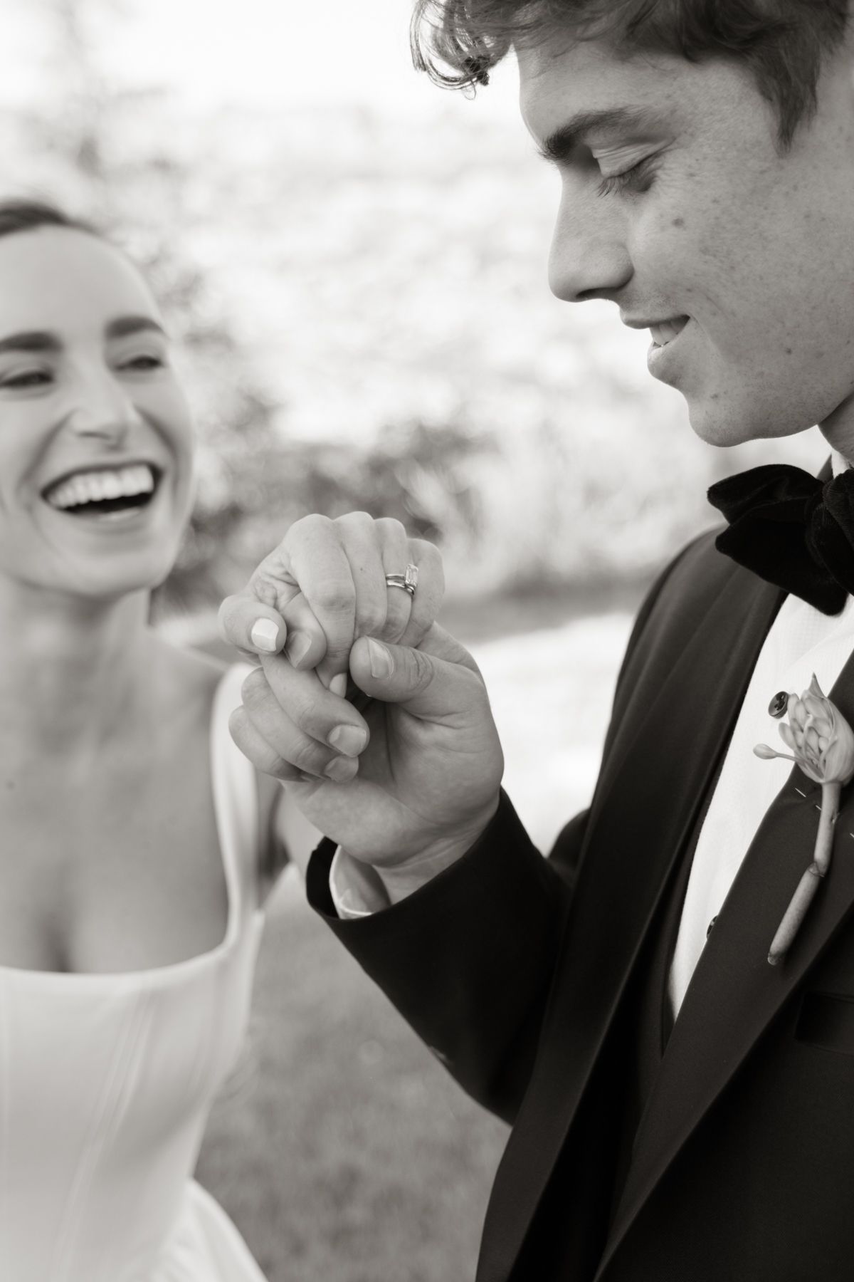 Black and white photo of a laughing bride and groom holding hands and admiring wedding rings outdoors.
