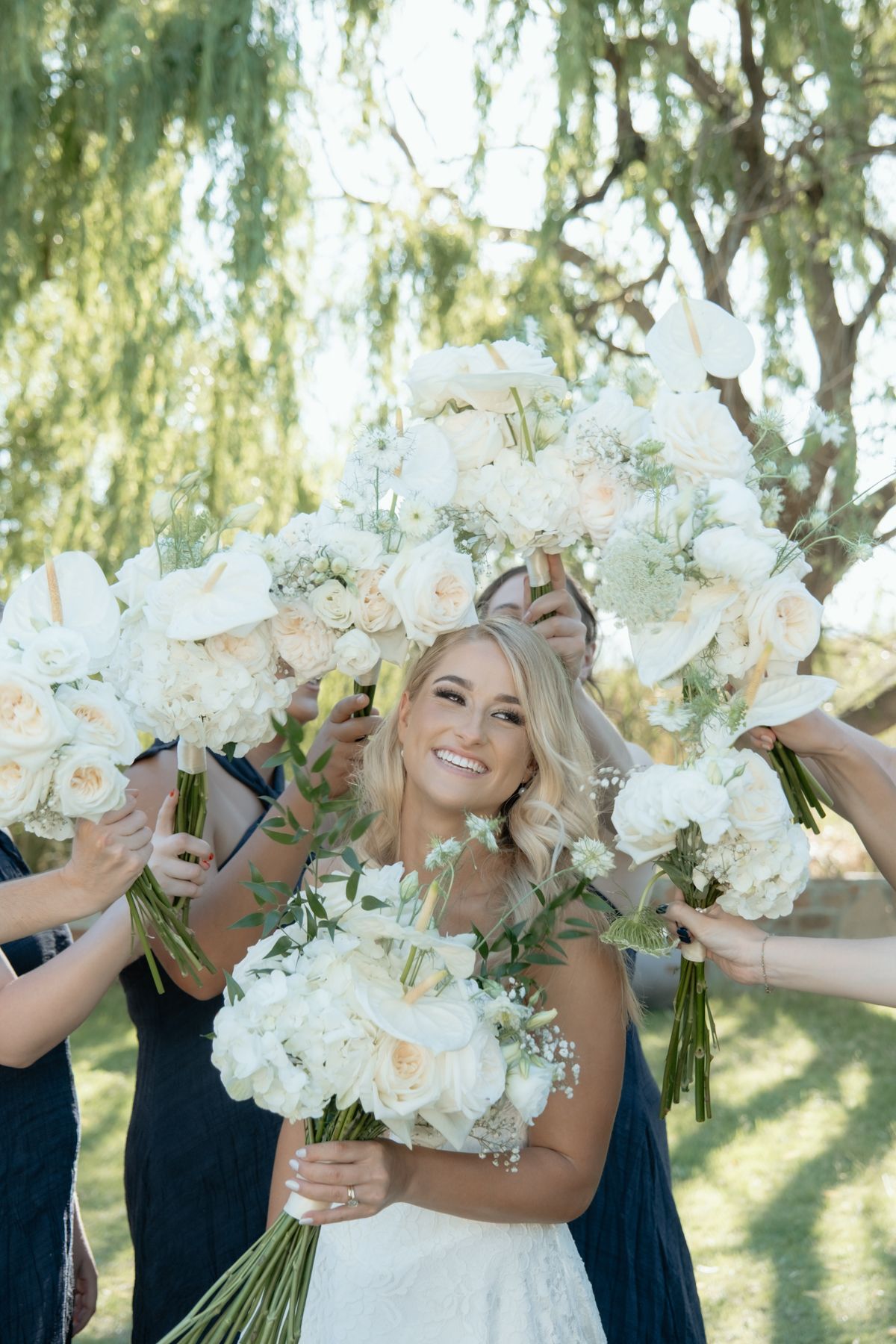 Smiling bride surrounded by white peony and rose bouquets held up by bridesmaids under a willow tree.