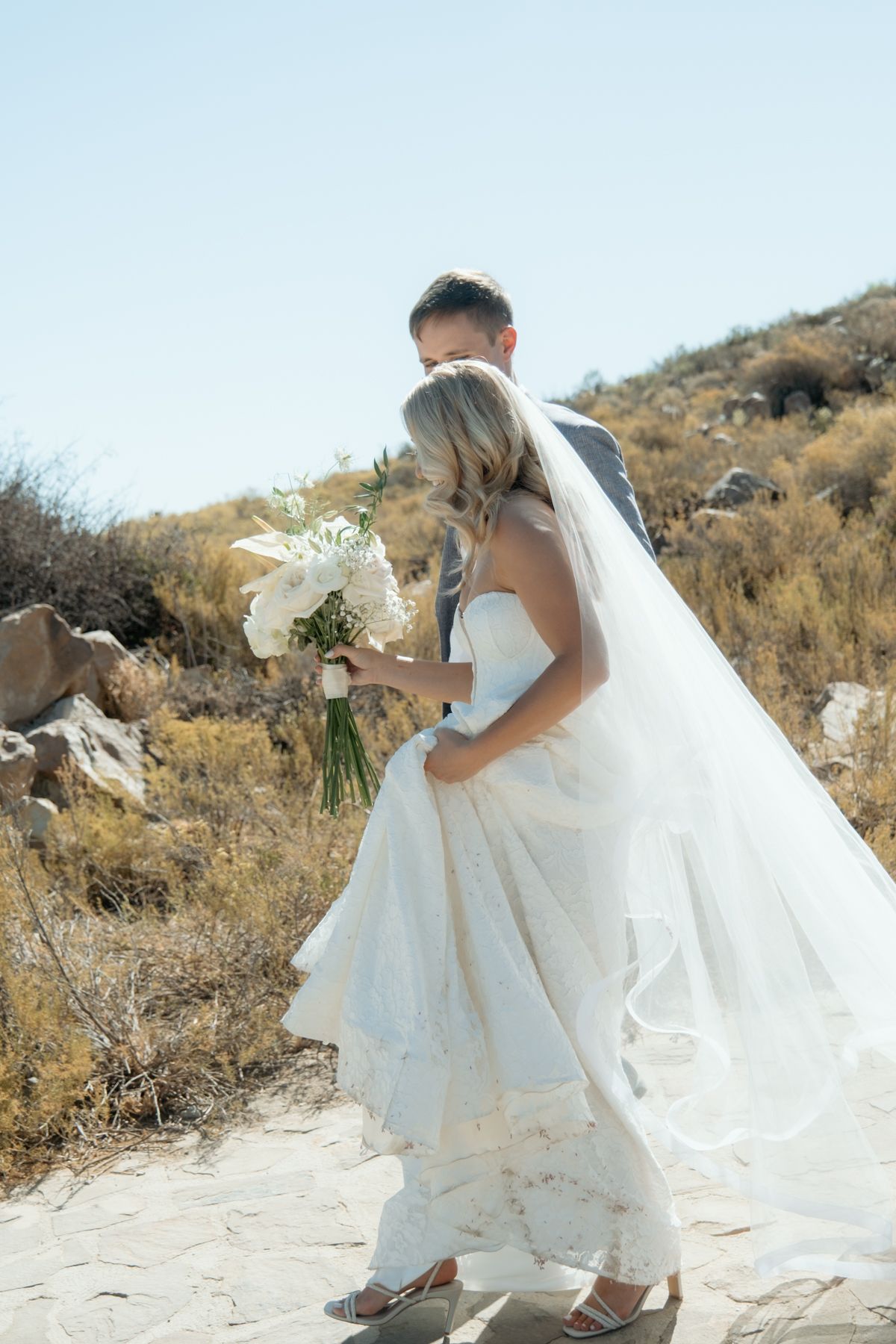 Bride and groom walking on stone path, bride holding white bouquet with veil flowing.