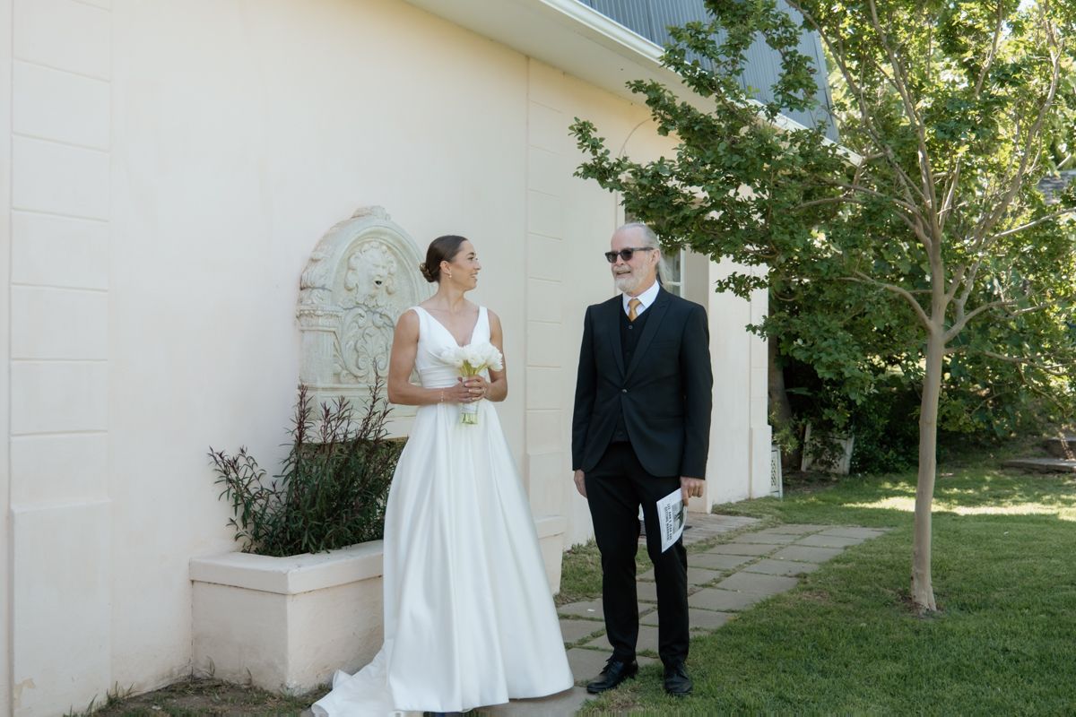 Bride in white gown stands with older gentleman beside a white wall in a sunny outdoor setting.