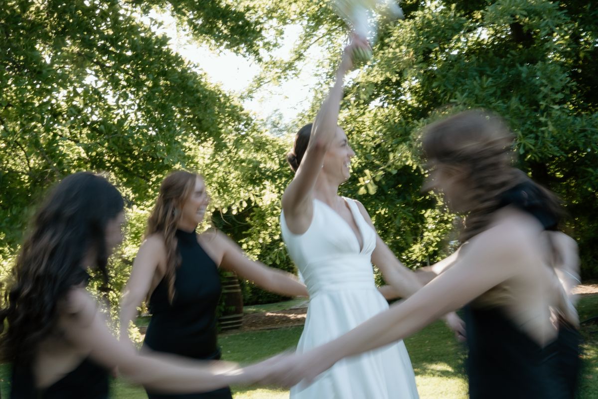 Bride in white dress dancing joyfully with bridesmaids in black under lush green trees in a garden setting.