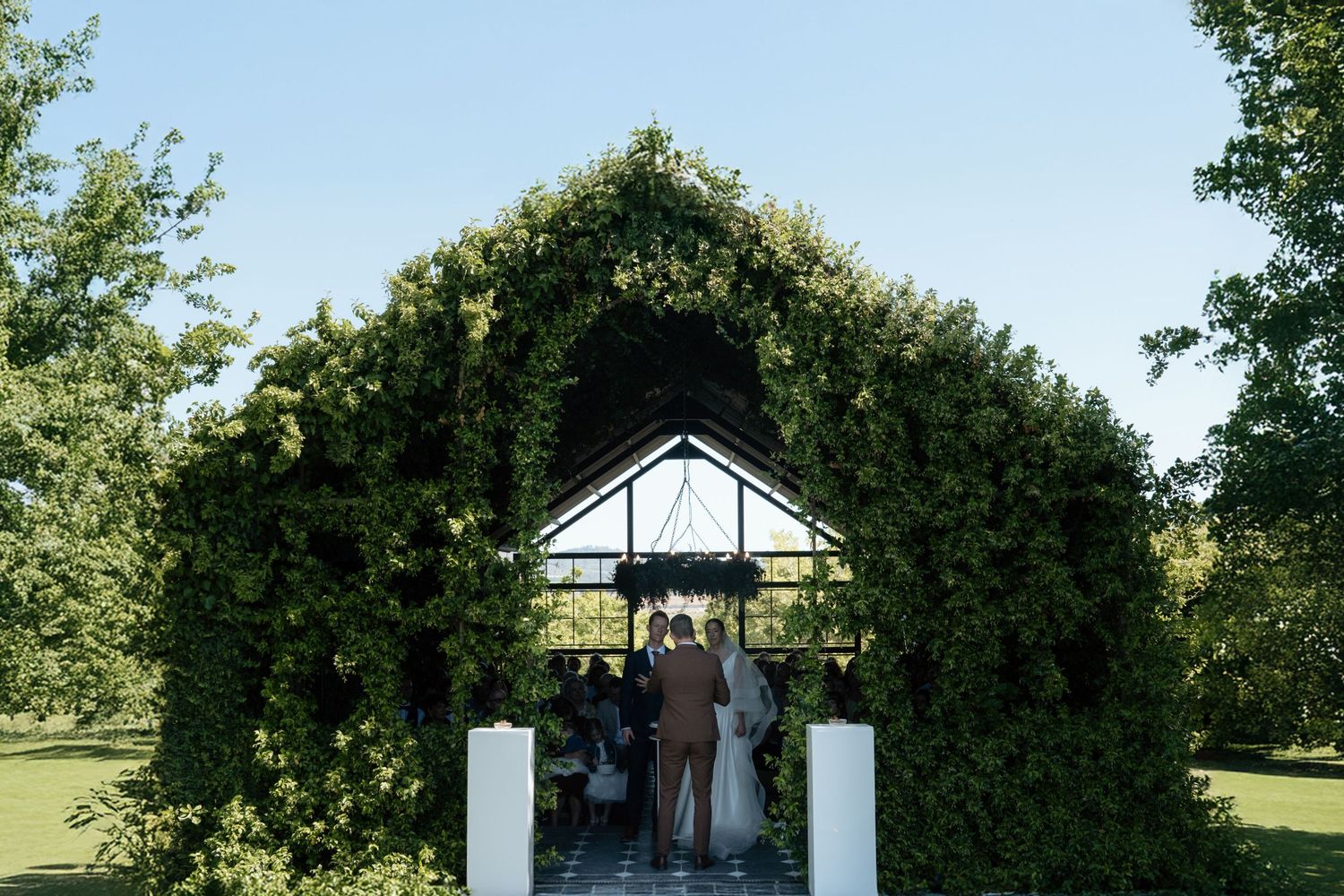 Couple stands beneath a lush green ivy-covered arch at an outdoor garden wedding ceremony on a sunny day.