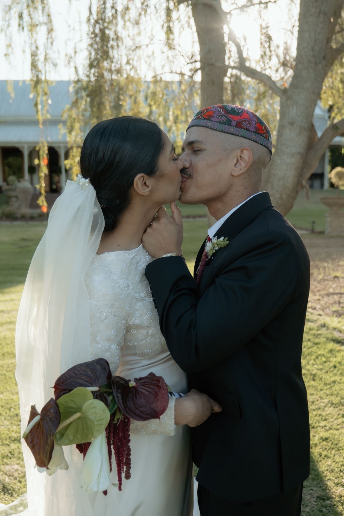 Bride and groom sharing first kiss outdoors with weeping willow tree in background.