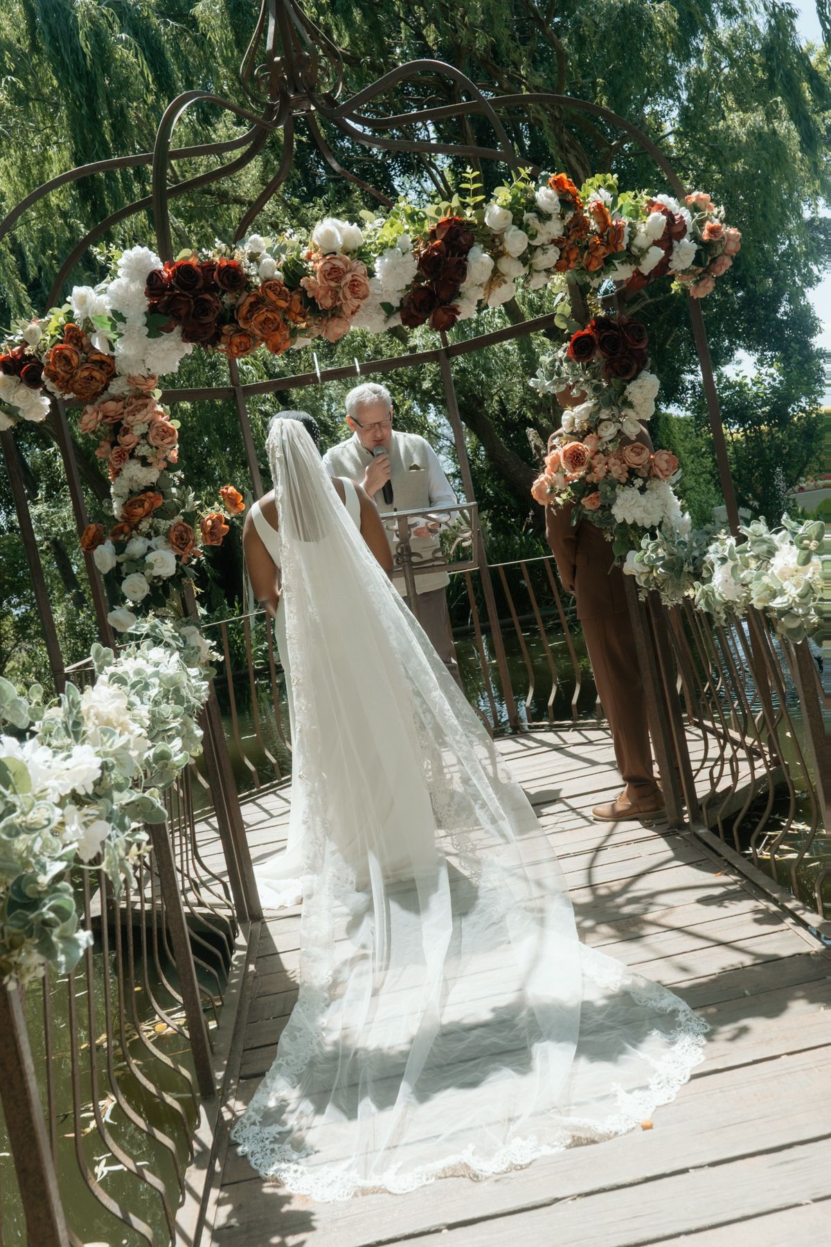 Bride in long lace veil stands at floral wedding arch gazebo with officiant.