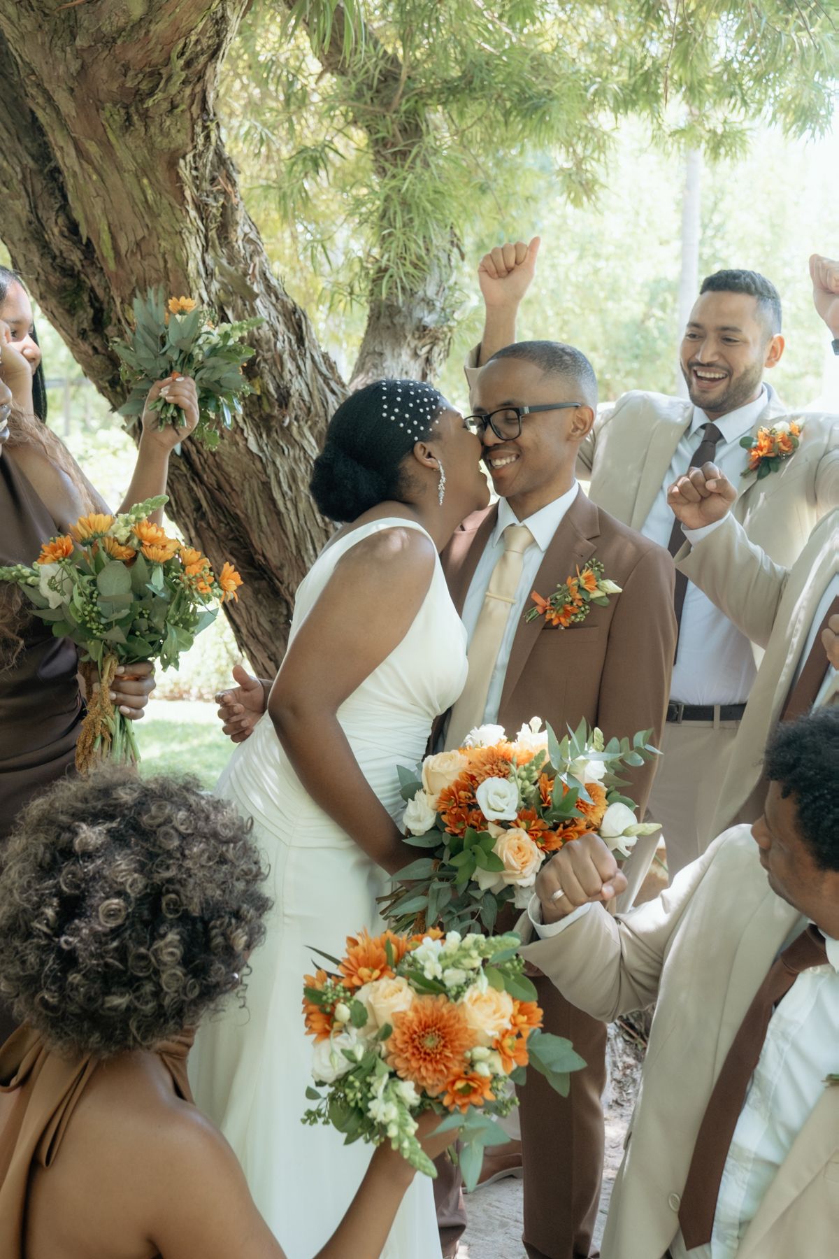 Joyful newlyweds share a kiss surrounded by their wedding party holding orange and white bouquets outdoors.