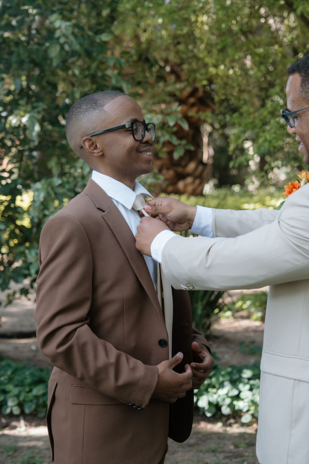 Groomsman in beige suit adjusts smiling grooms tie outdoors in lush green garden setting.