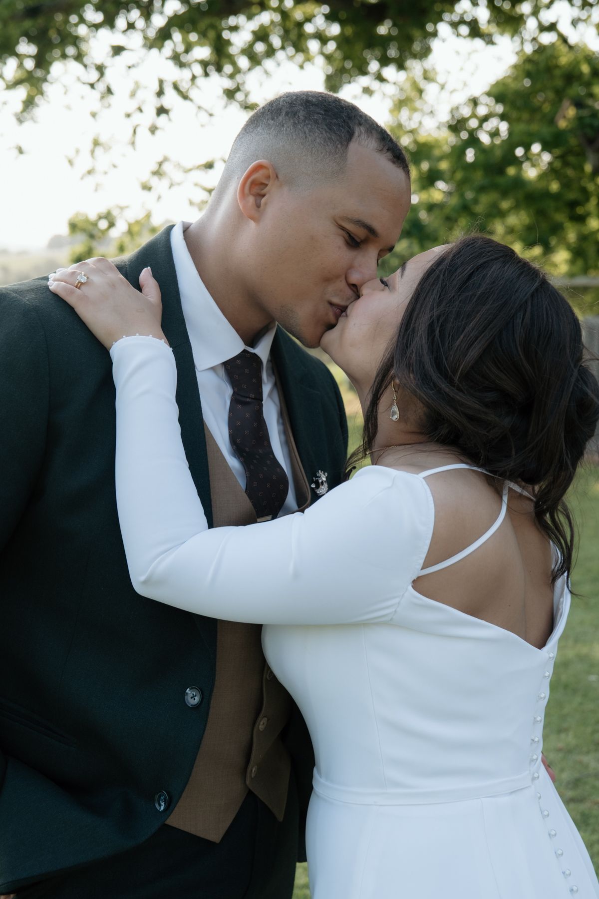 A bride and groom share a romantic kiss outdoors, surrounded by green trees at their wedding celebration.