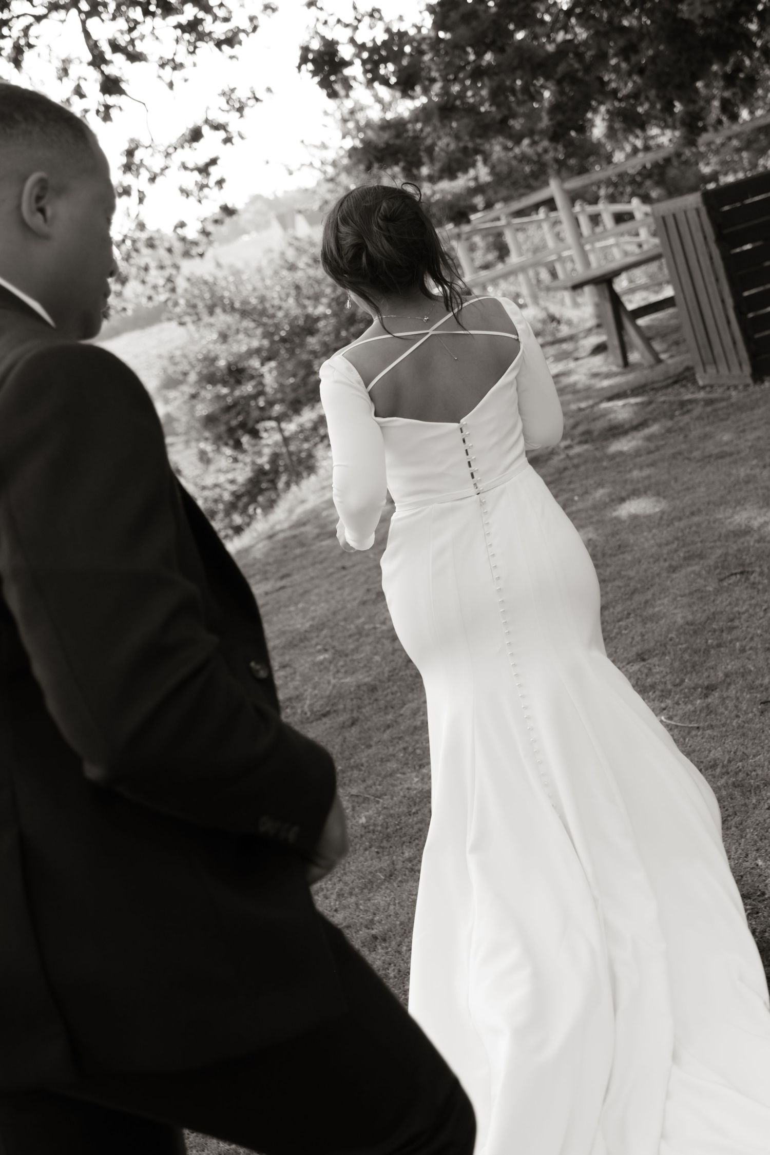 Black and white photo of bride in elegant backless gown walking with groom outdoors.