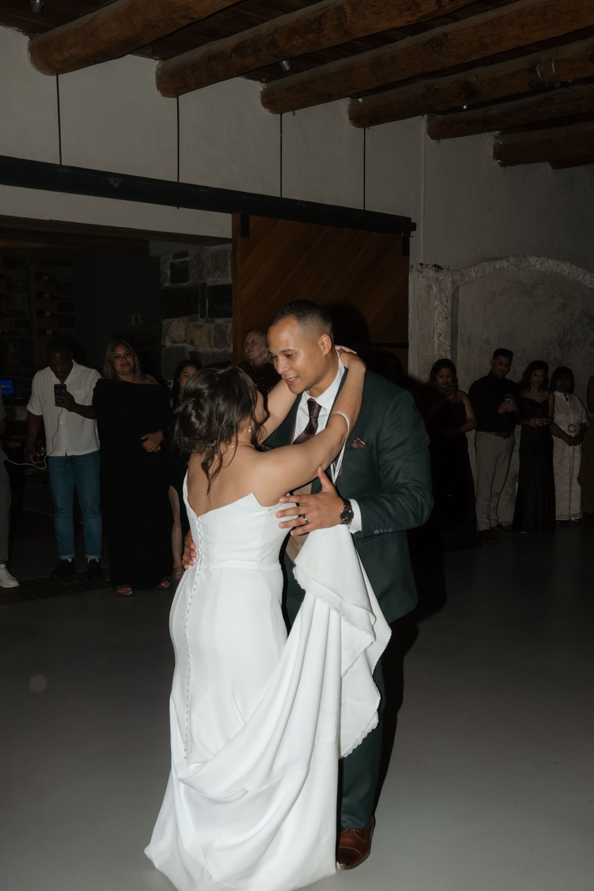 A bride and groom share their first dance in a rustic venue surrounded by wedding guests watching joyfully.