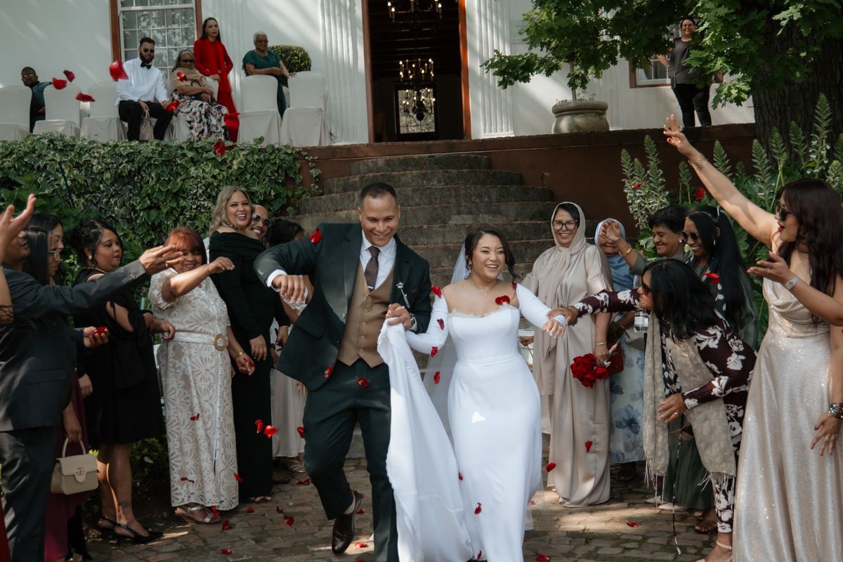 Joyful newlyweds walk through cheering guests throwing confetti outside ceremony venue in sunny garden.